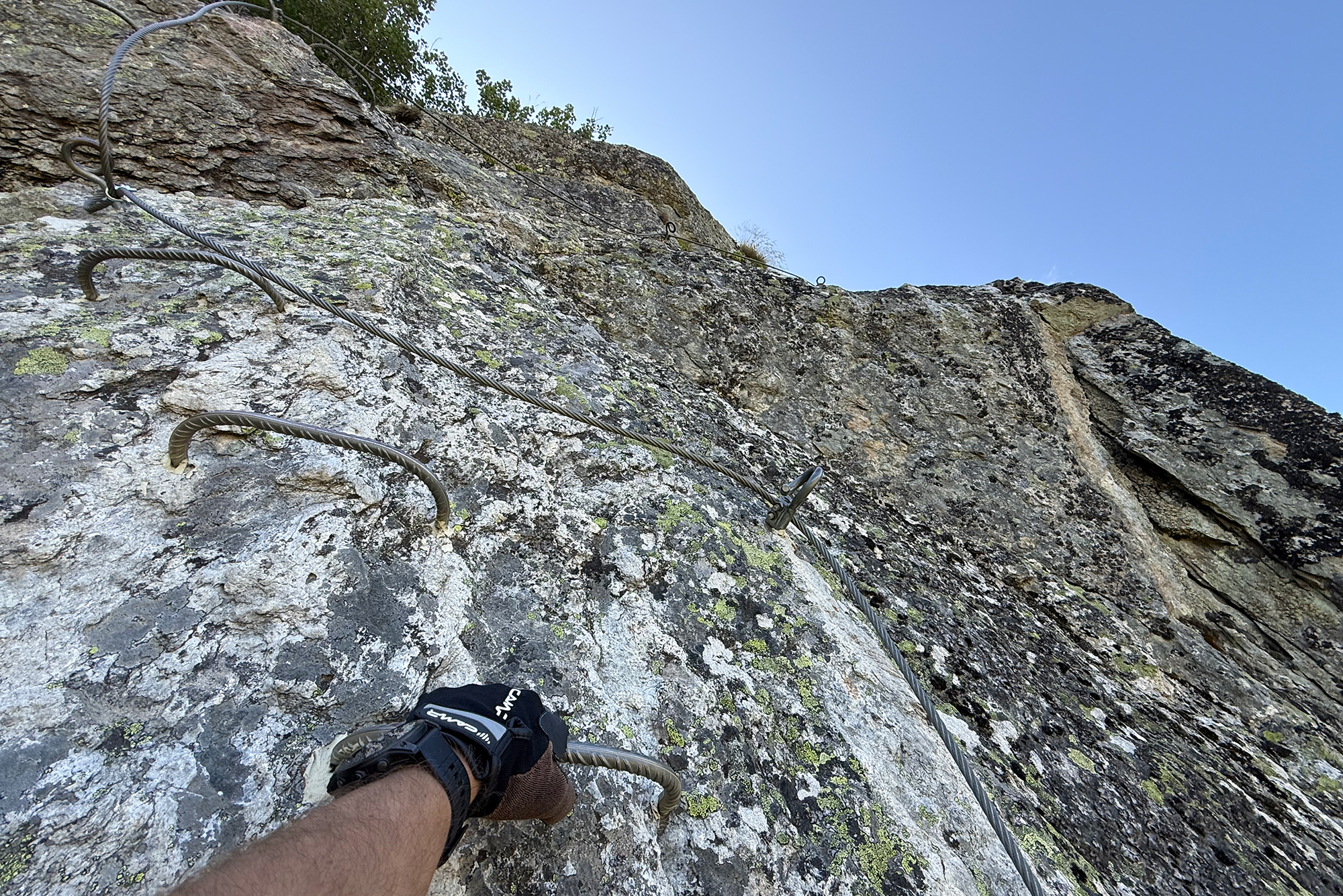 Rocky cliff with metal safety cables running along the route; a climber's gloved hand grips a cable.