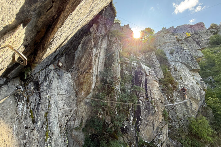 Cliffside via ferrata with steel cables and rungs along a rocky wall under bright sun; a hiker stands on a narrow metal bridge.