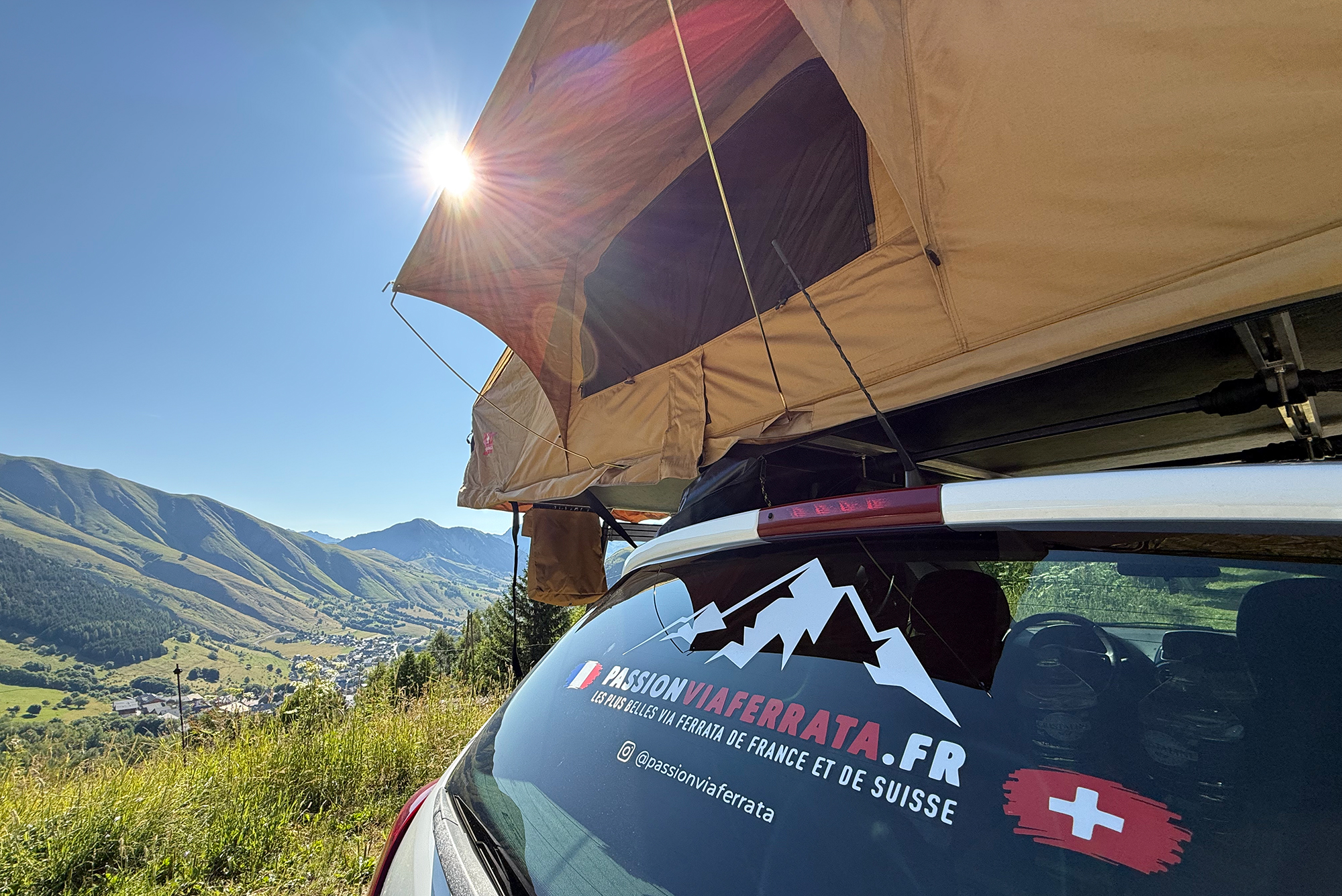SUV with a tan roof-top tent mounted, sun glare over a clear blue sky and alpine valley in the background