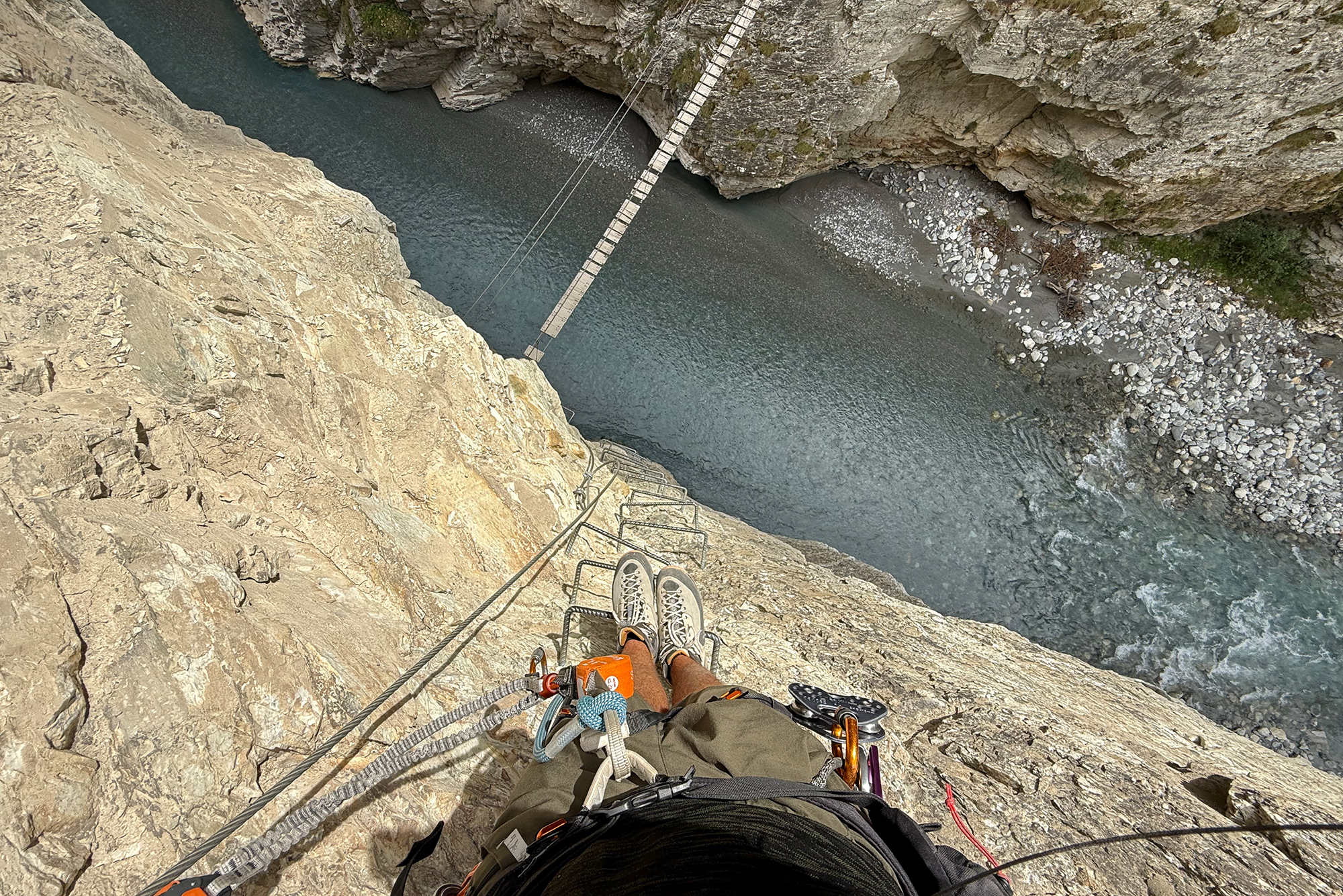 Climber wearing gear rappelling down a steep rocky cliff beside a river with a rope bridge overhead and harness equipment visible below.