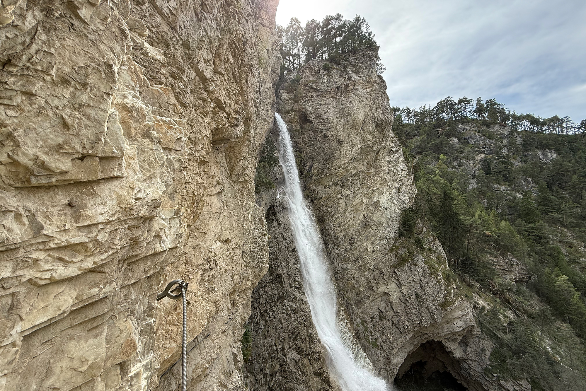 Waterfall cascading down a steep rocky cliff with a metal handrail in the foreground and forested hills beyond under a cloudy sky.