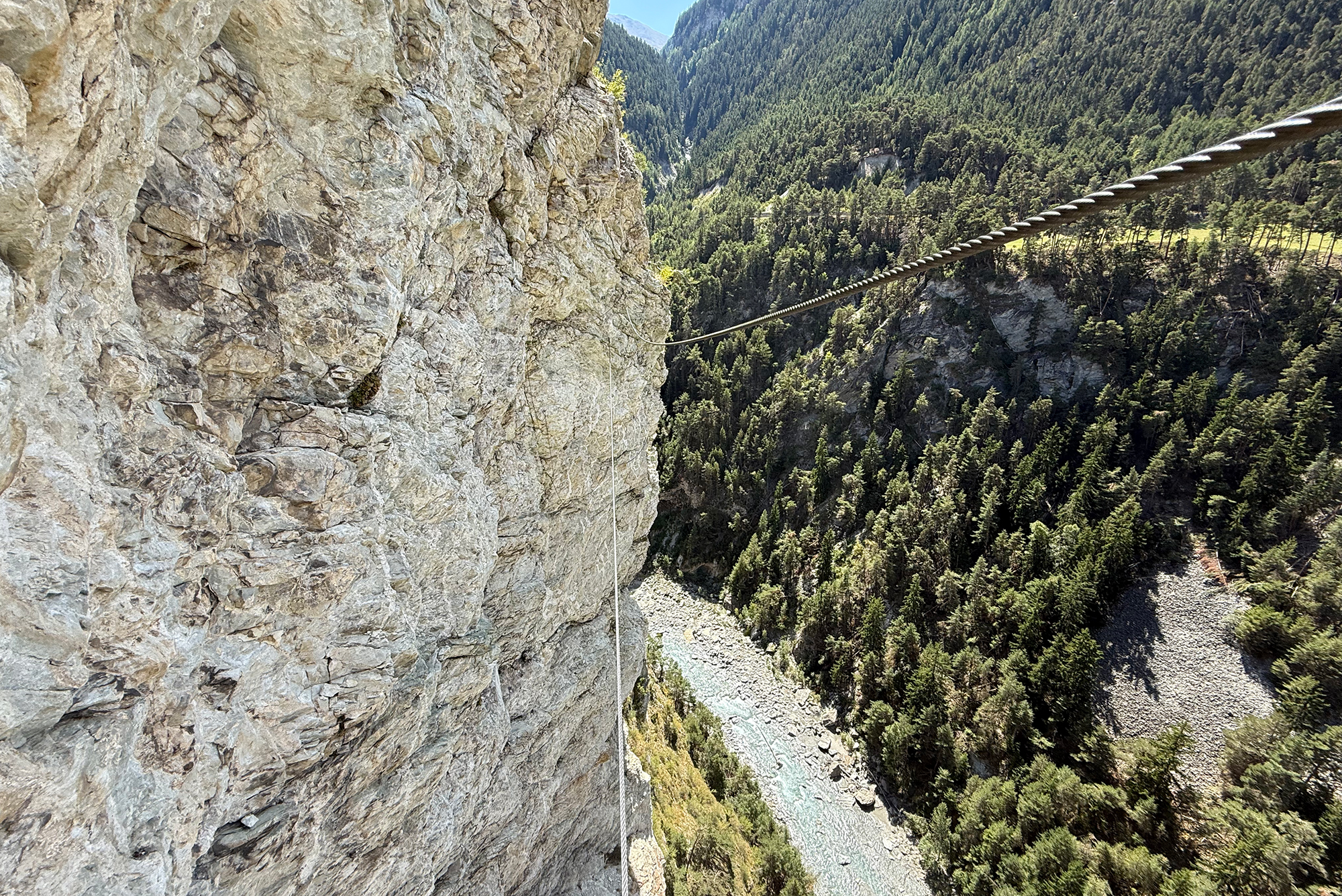 Steep rocky cliff with a rope line across a deep, forested valley below two rock faces.