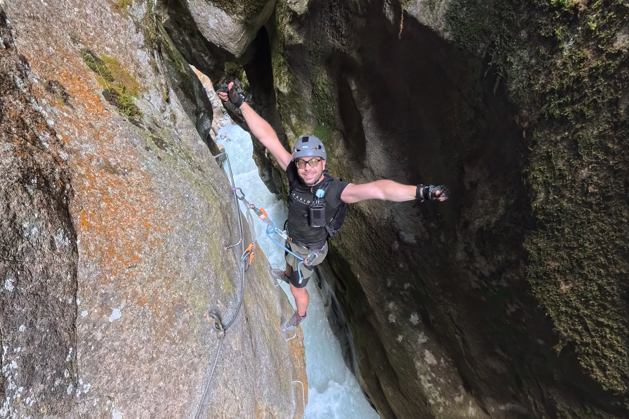 Person wearing a helmet and safety gear traversing a narrow rock canyon with ropes and metal anchors above a rushing pool of water.