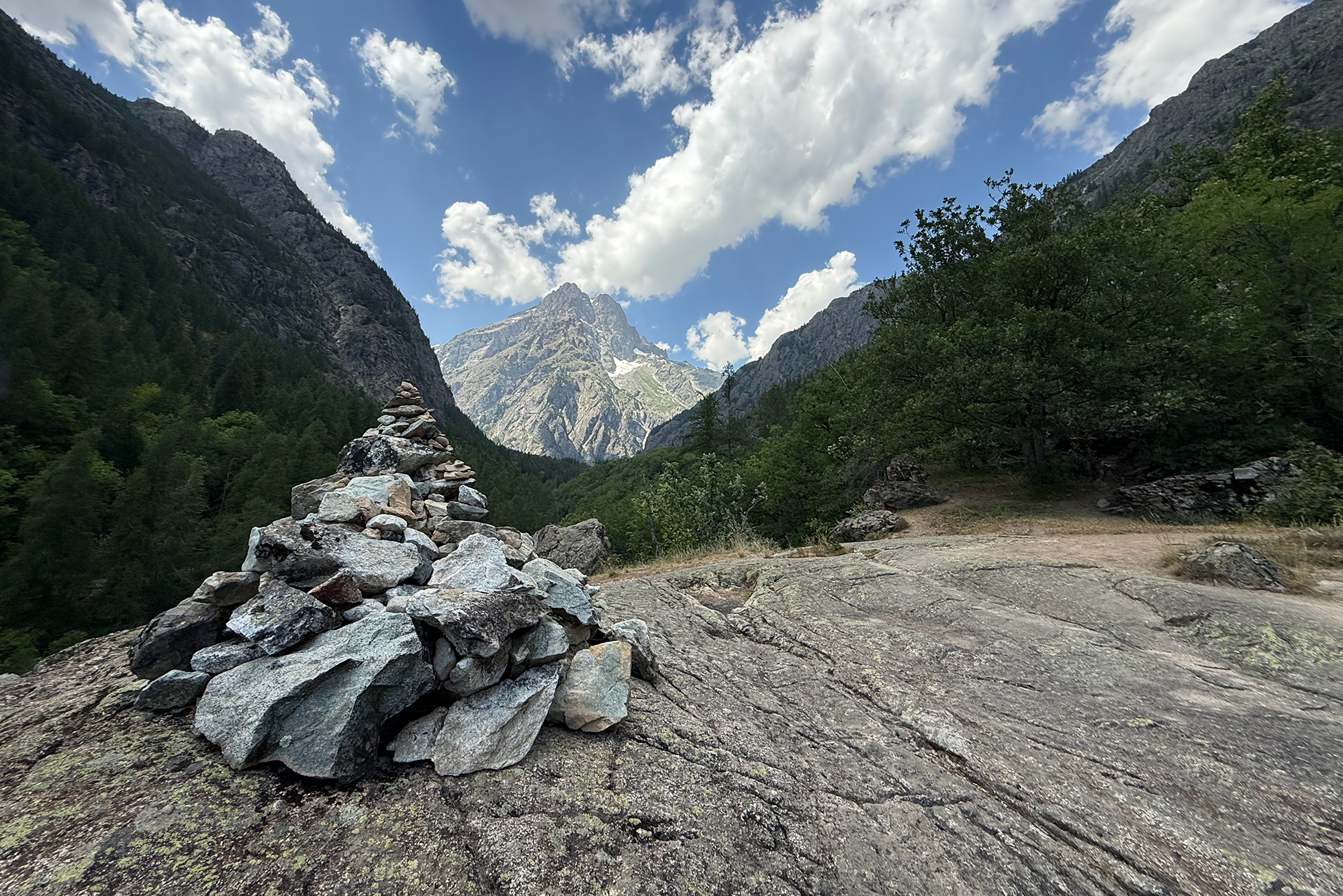 Rock cairn on a cracked rocky foreground with pine-covered slopes and jagged mountains under a blue cloudy sky