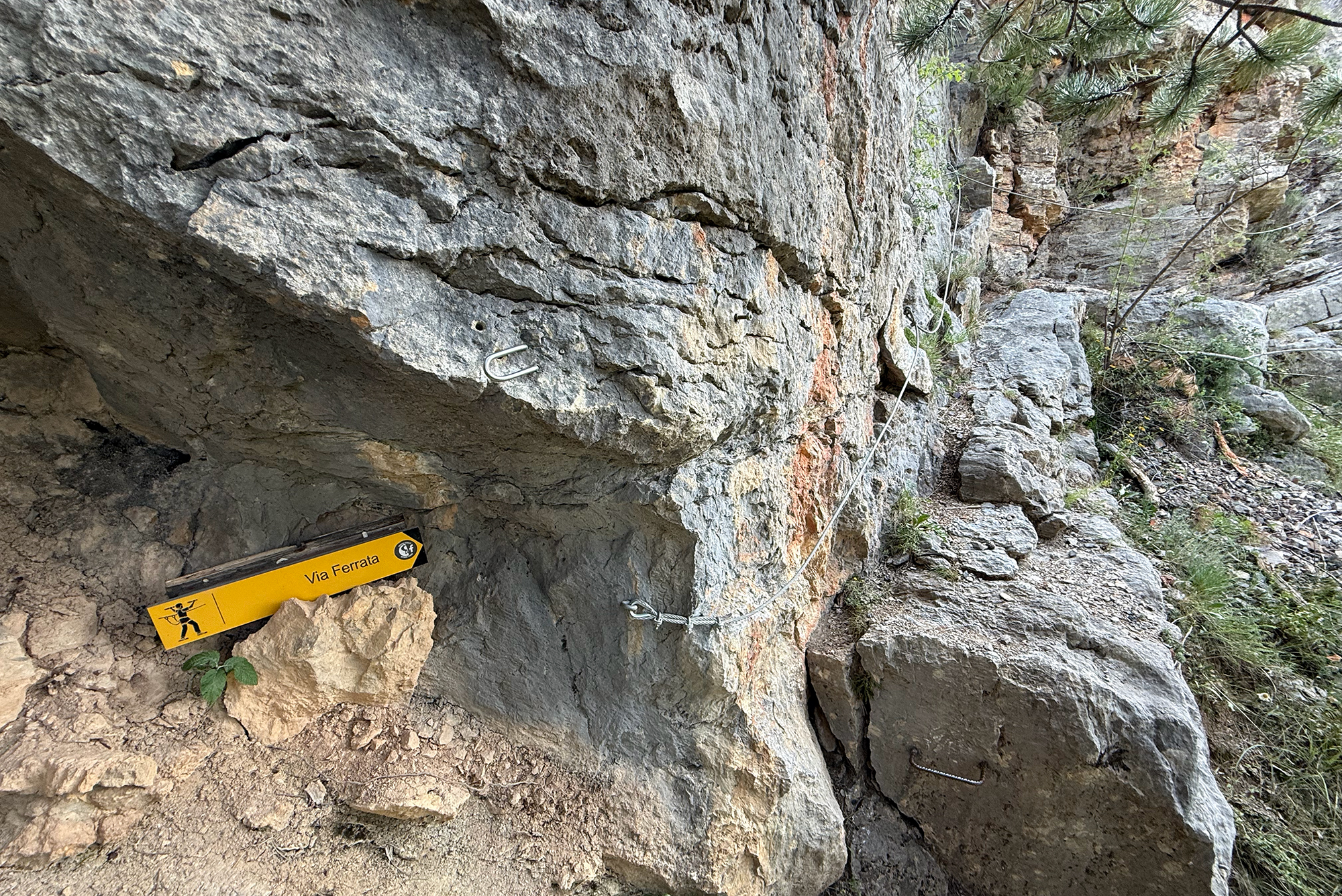 Rocky cliff face with safety cables anchored along the rock and a yellow Via Ferrata sign indicating a climbing route.