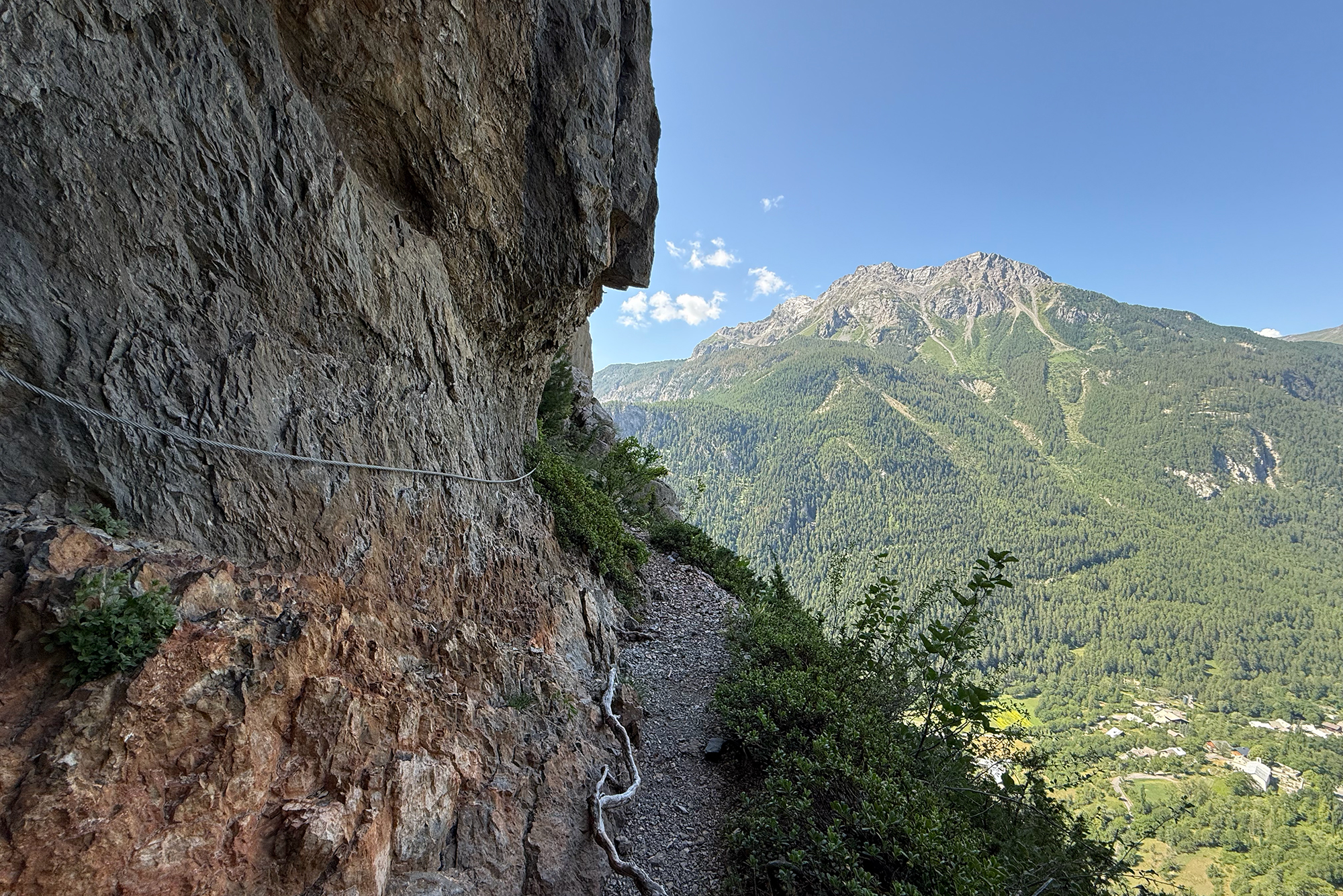 Rocky mountainside trail hugging a sheer cliff, overlooking a forested valley and distant village under a blue sky.
