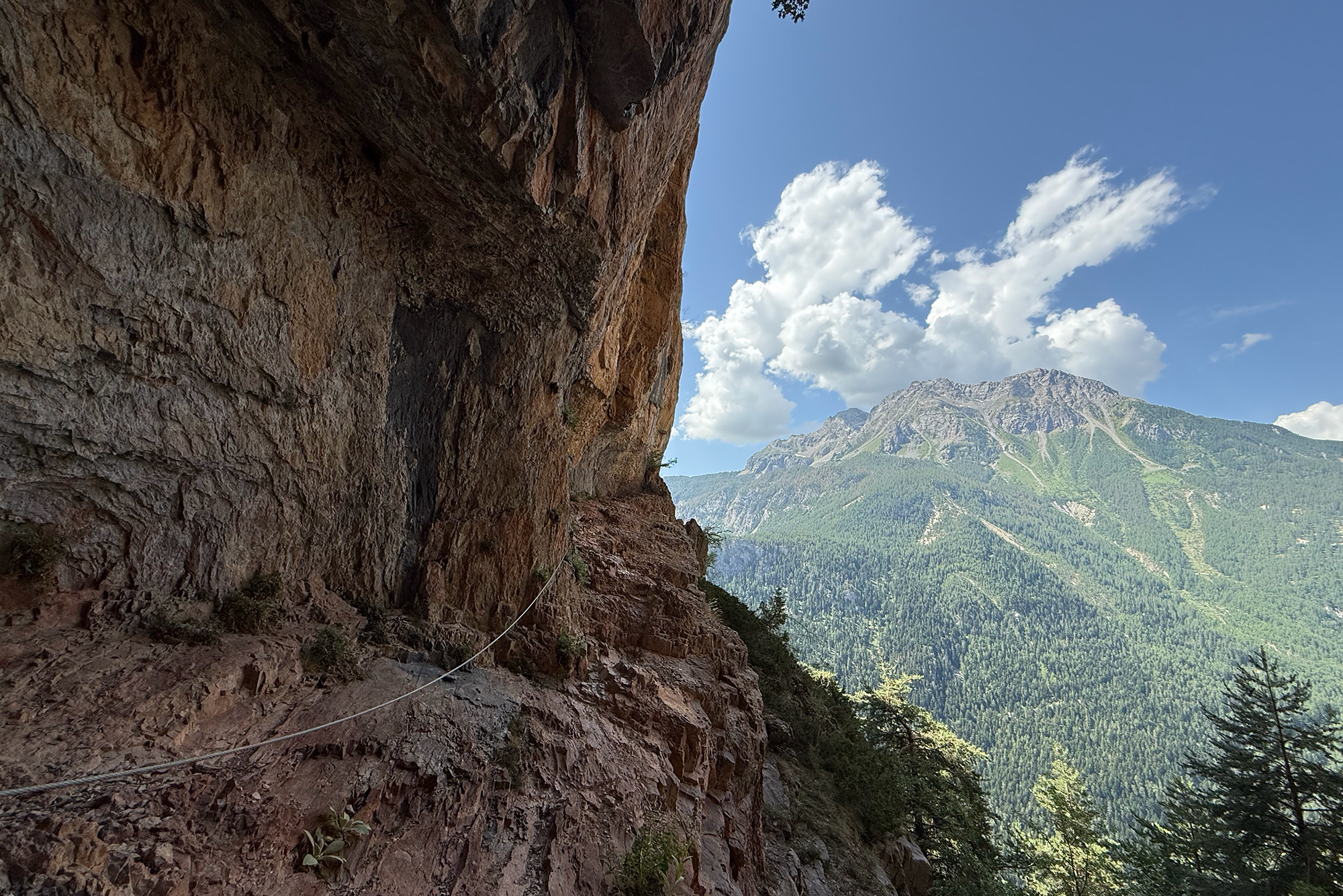 Rugged rocky cliffside along a mountain trail with a rope, overlooking a forested valley and distant peaks under a blue sky.