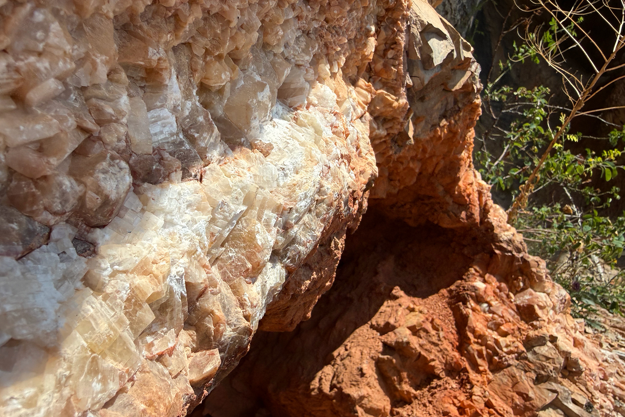 Rugged rock face with beige and white crystalline minerals, orange soil, and sparse green vegetation along the edge.