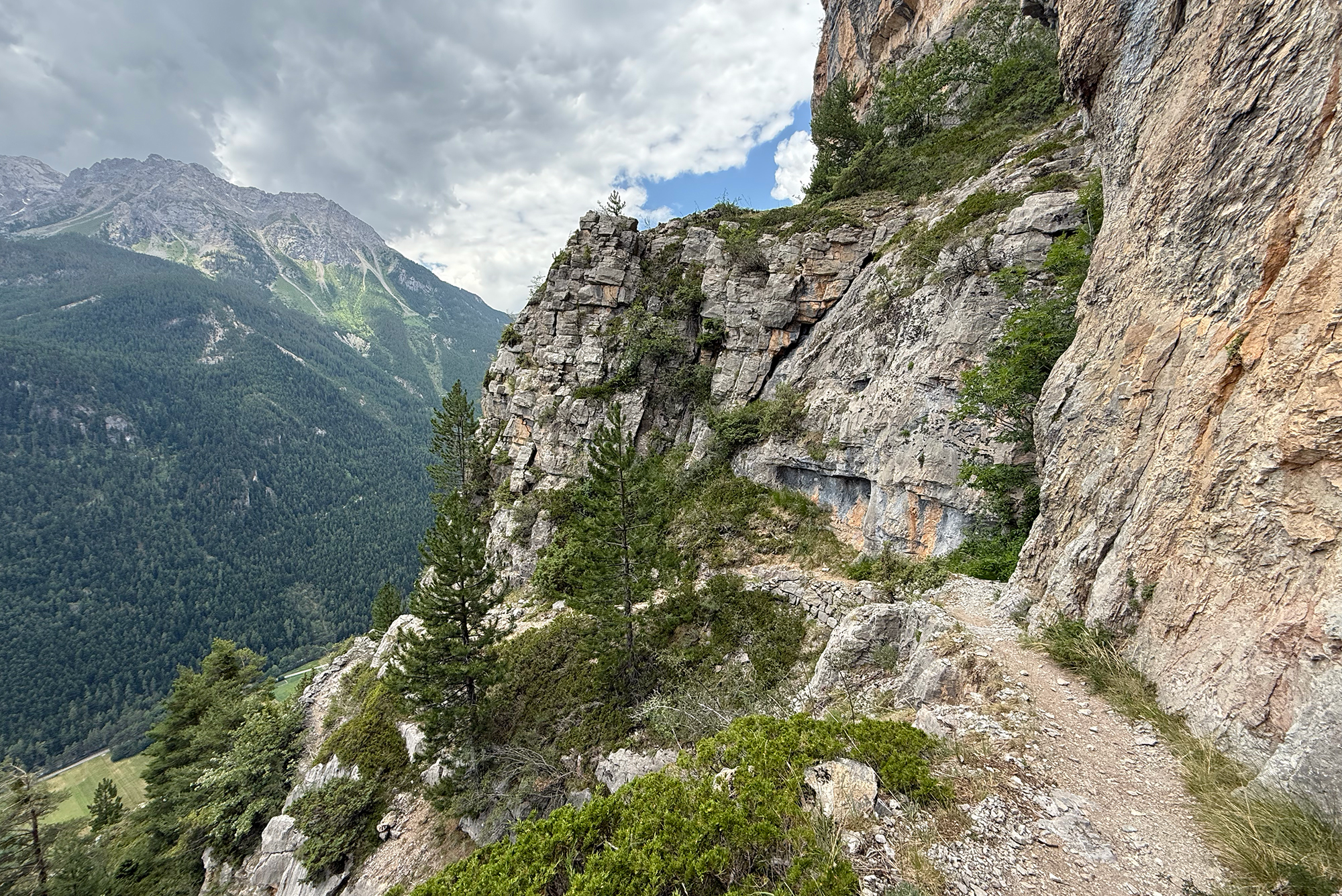 Steep rocky cliff face with sparse vegetation along a mountain path, overlooking a forested valley and distant snow-free peaks under a cloudy sky.