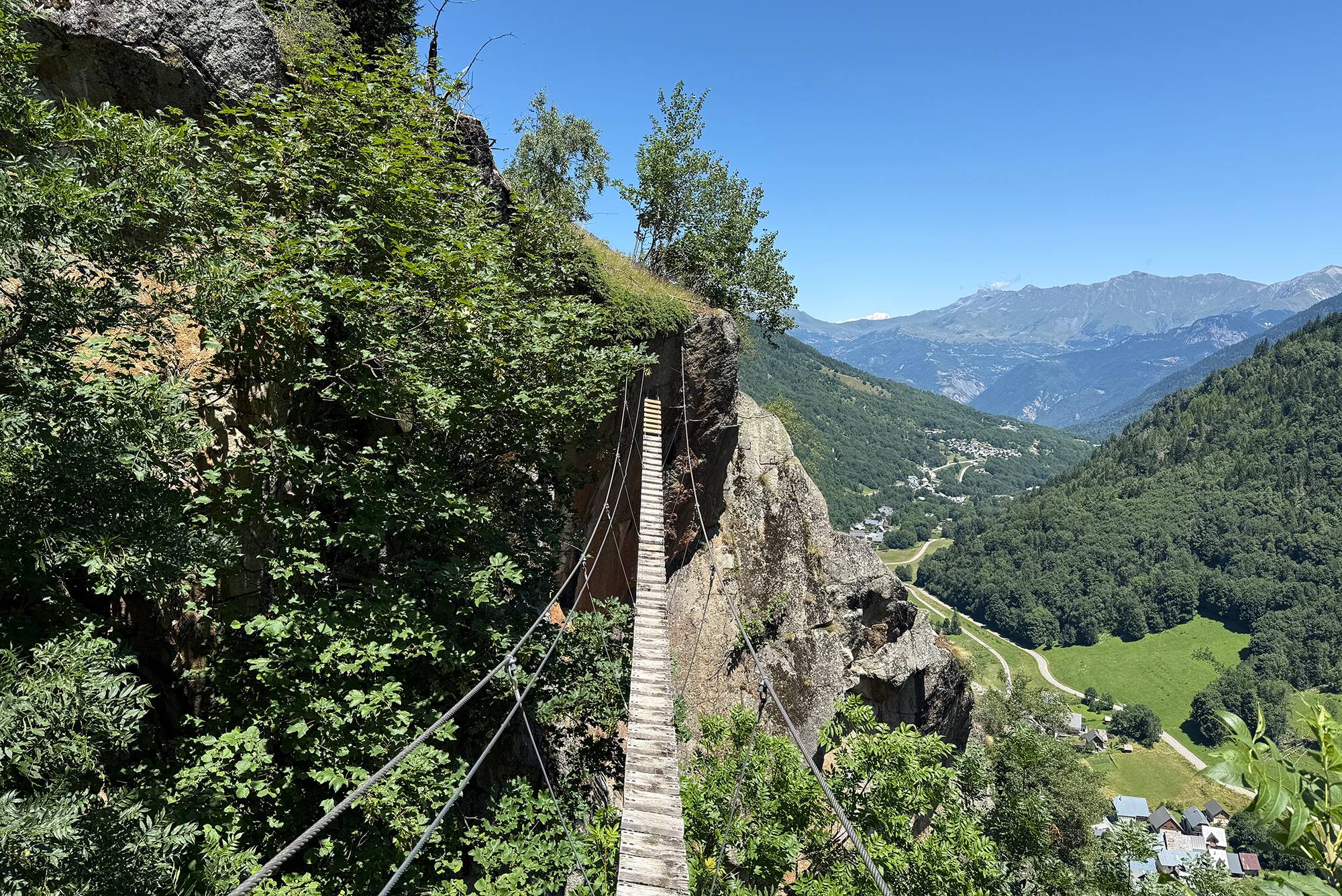 Wooden suspension footbridge along a rocky cliff, with dense greenery and mountainous valley below under a clear blue sky