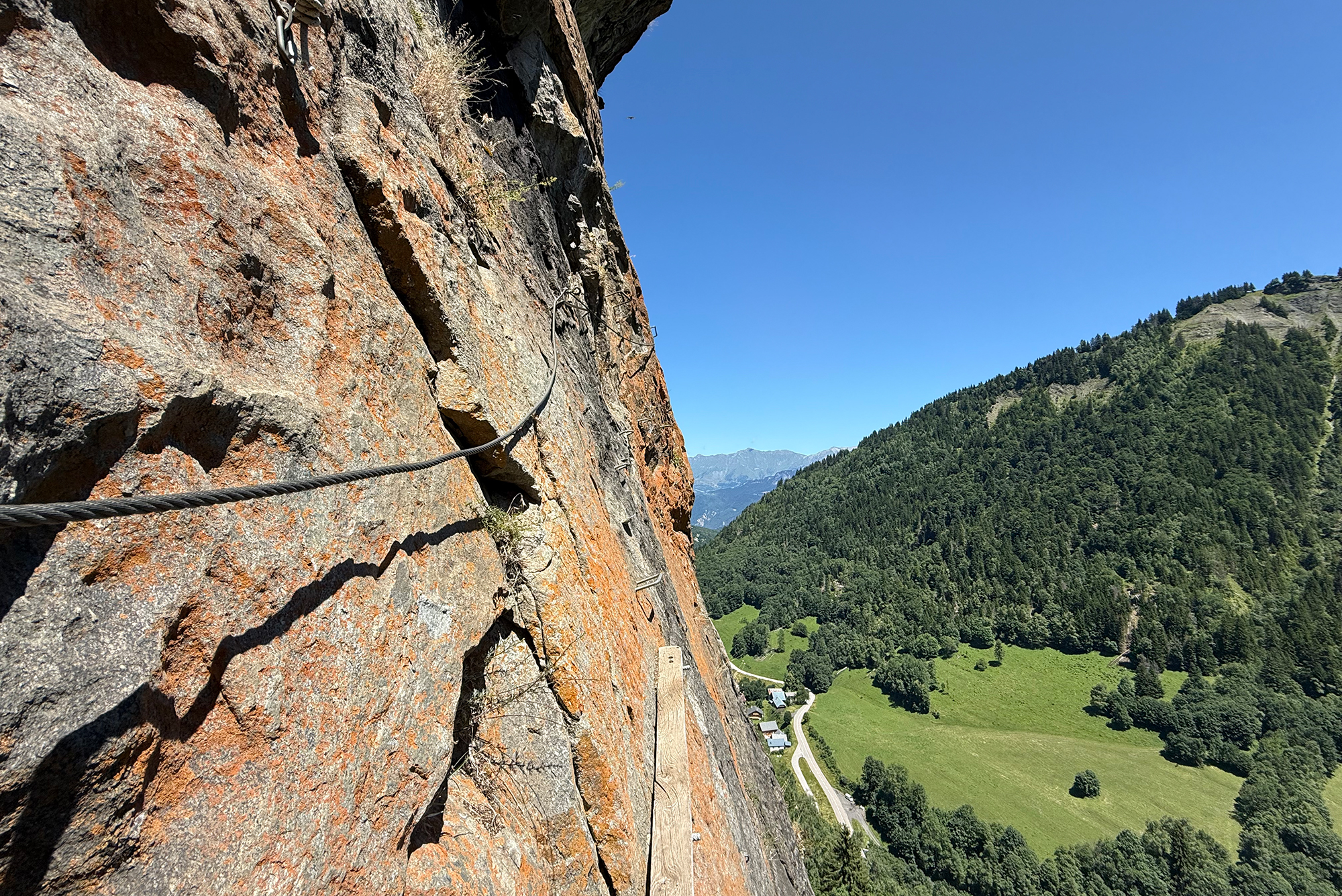 Rock cliff with metal climbing cables and safety ladders on the left, overlooking a green valley and distant mountains under a clear blue sky.