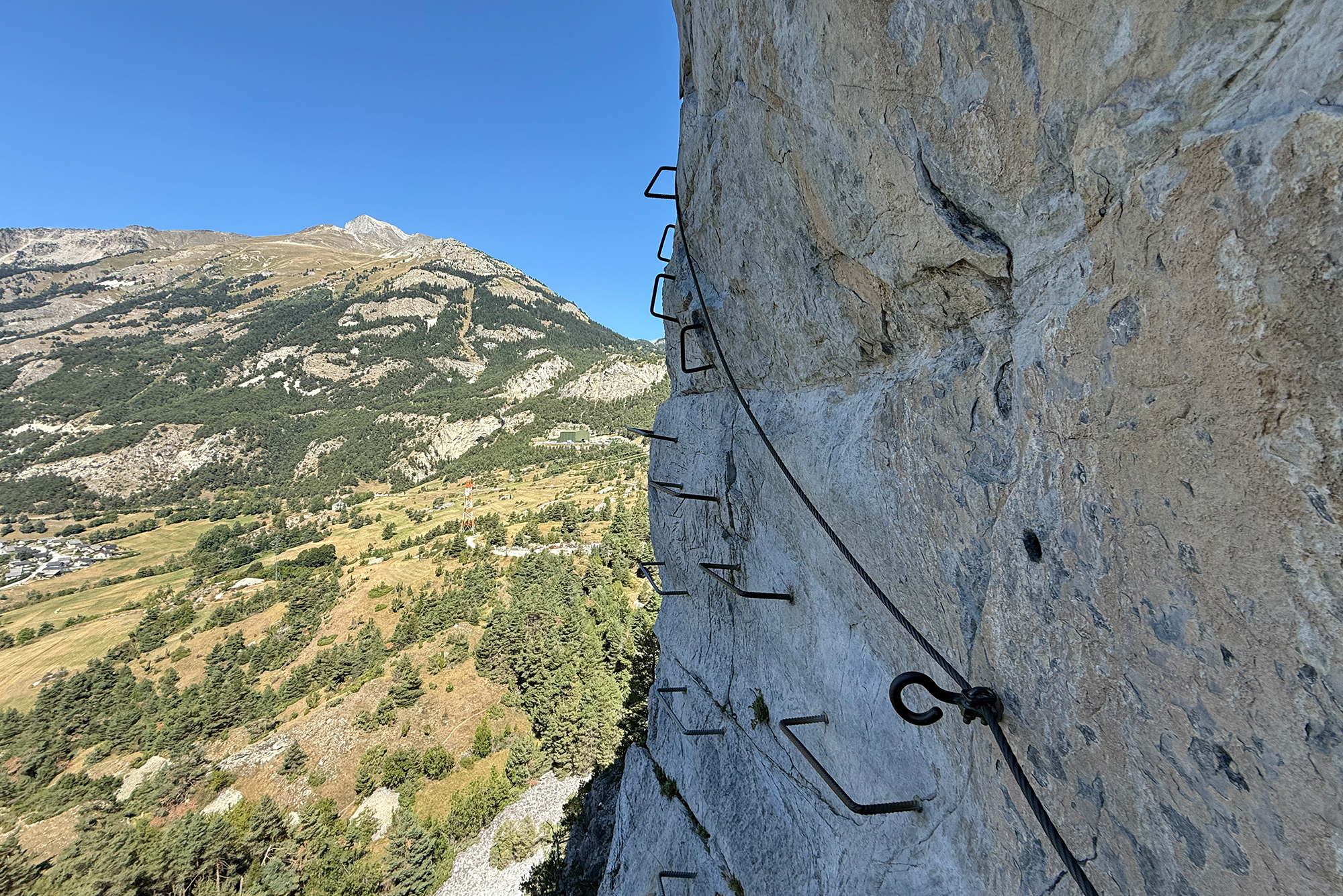Rock cliff side with anchored metal rungs and a safety rope, overlooking a valley and distant mountains under a clear blue sky.