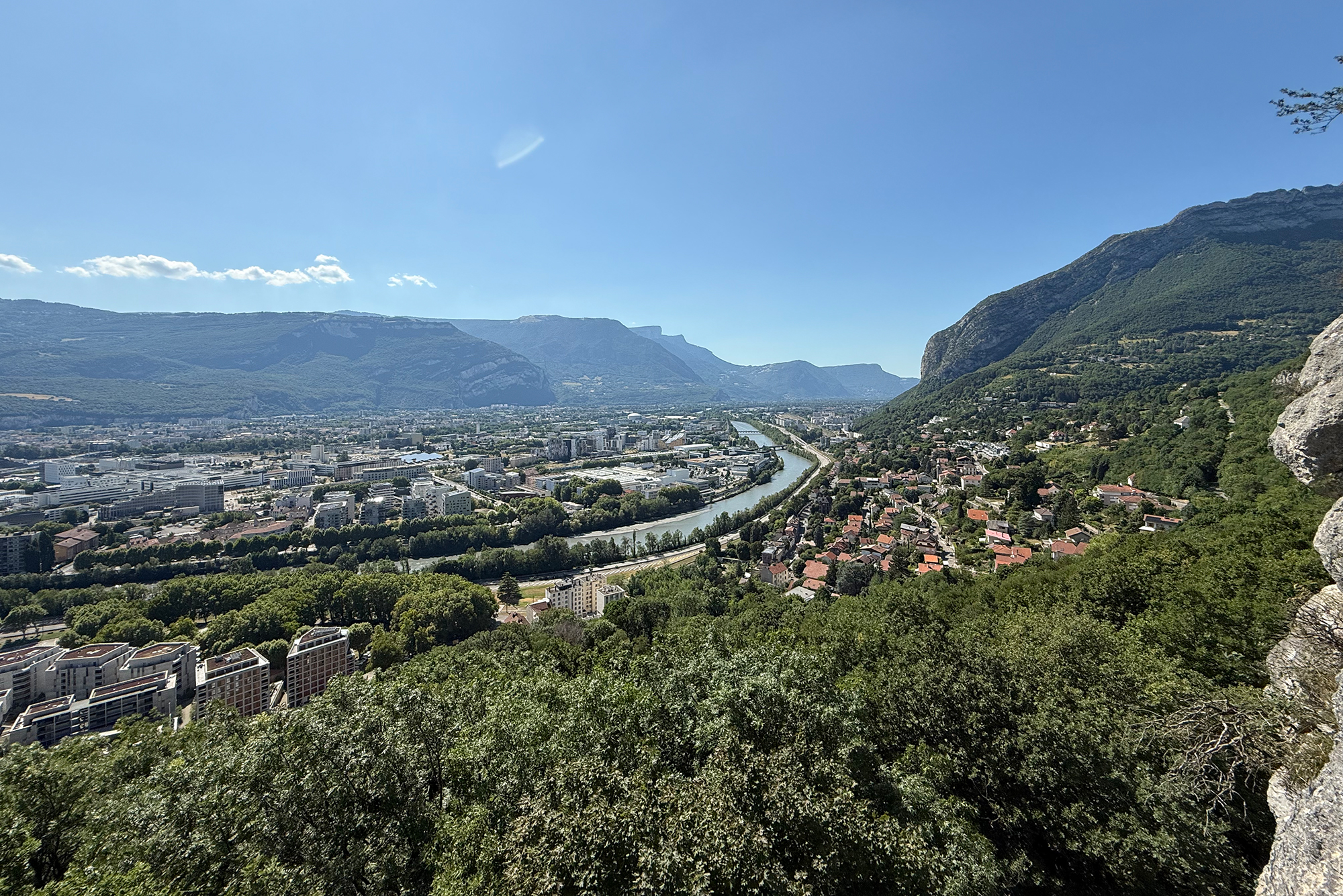 Aerial view of a city in a valley with a winding river, flanked by green hills and distant mountains under a clear blue sky.