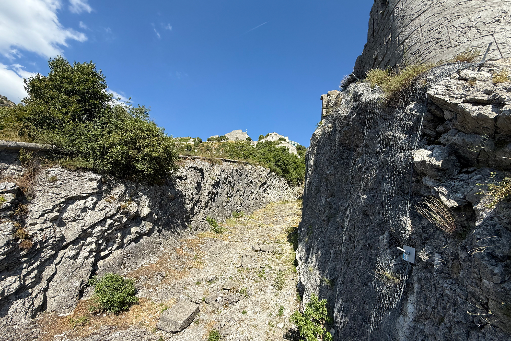 Narrow path between ancient stone walls with a distant hillside fortress under a blue sky.