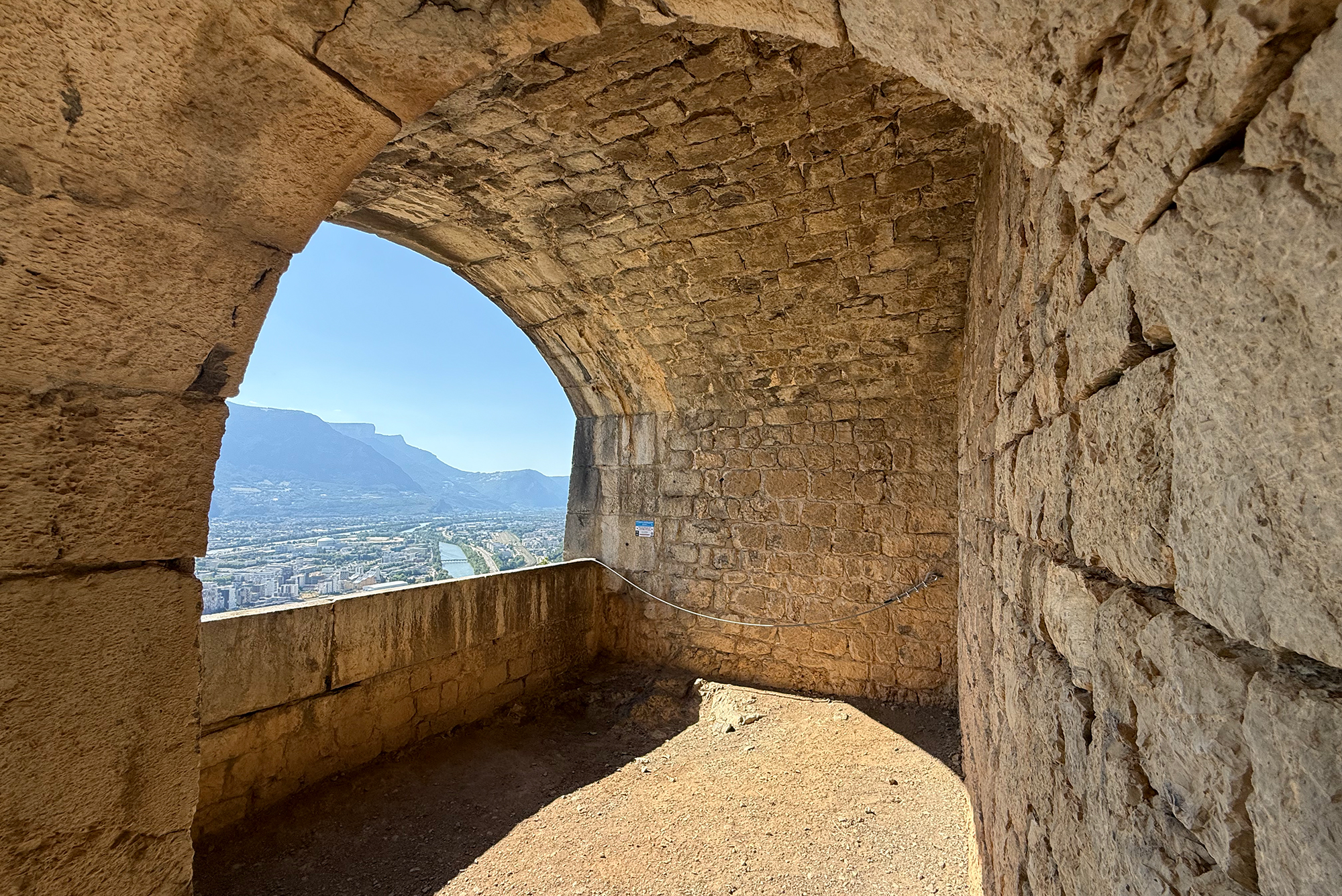 Stone stone-arched fortress passage with a view of a city and distant mountains beyond the opening.