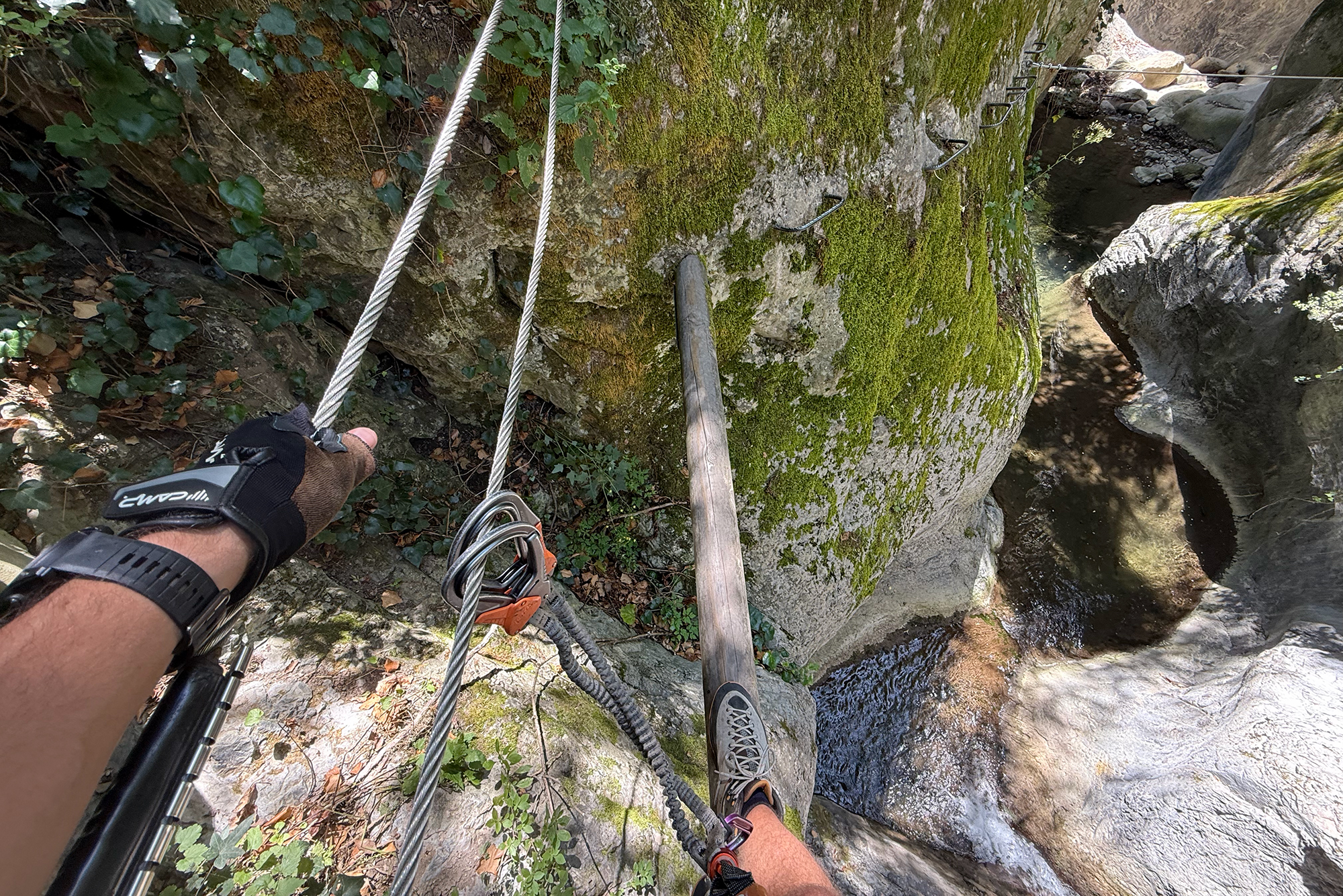 Climber's gloved hand grips a steel cable on a mossy rock cliff, with carabiners and harness visible. Perspective shot along a via ferrata route.