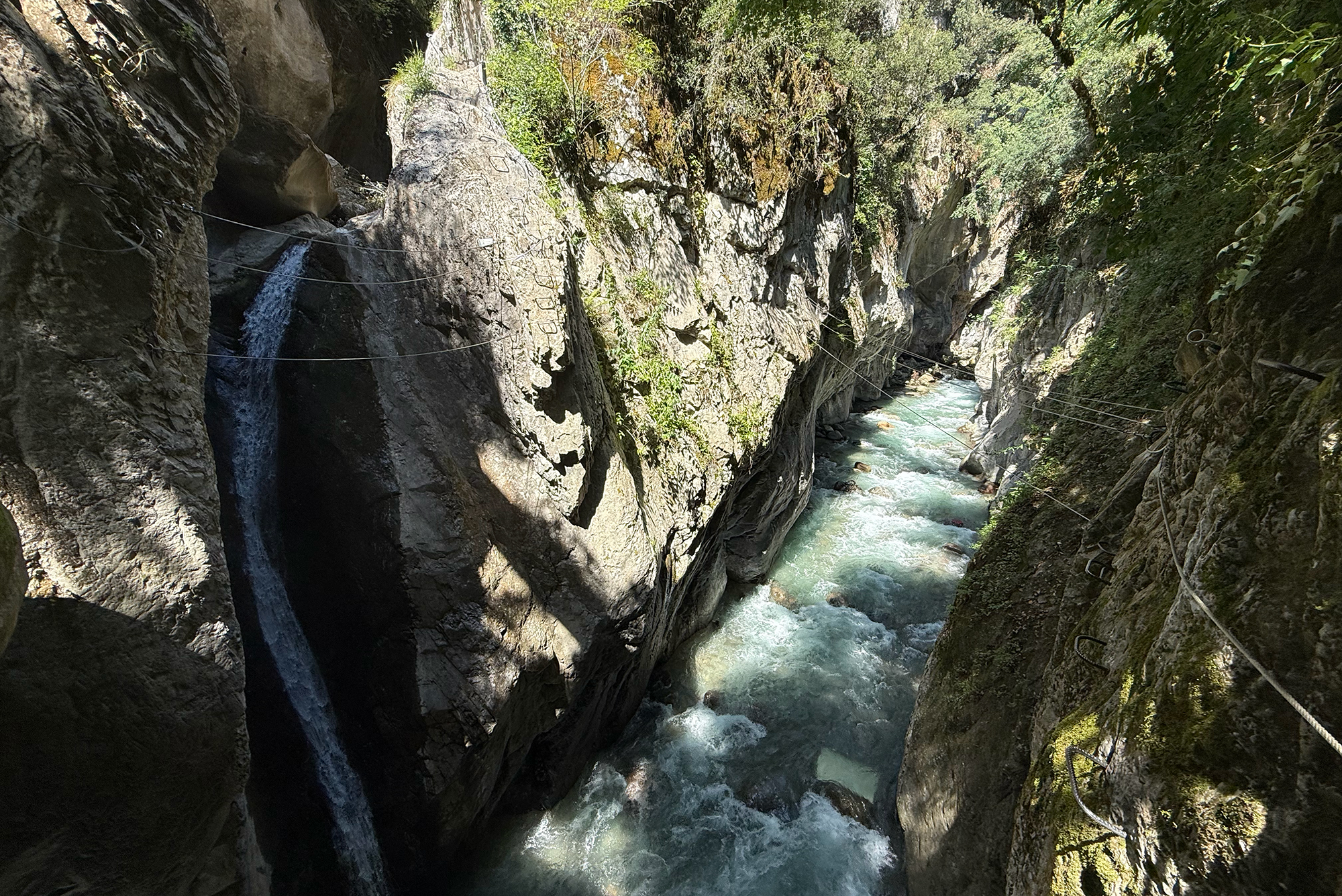 Narrow rocky gorge with steep, mossy walls and a fast-flowing turquoise river at the bottom, with metal cables along the cliff for traversal