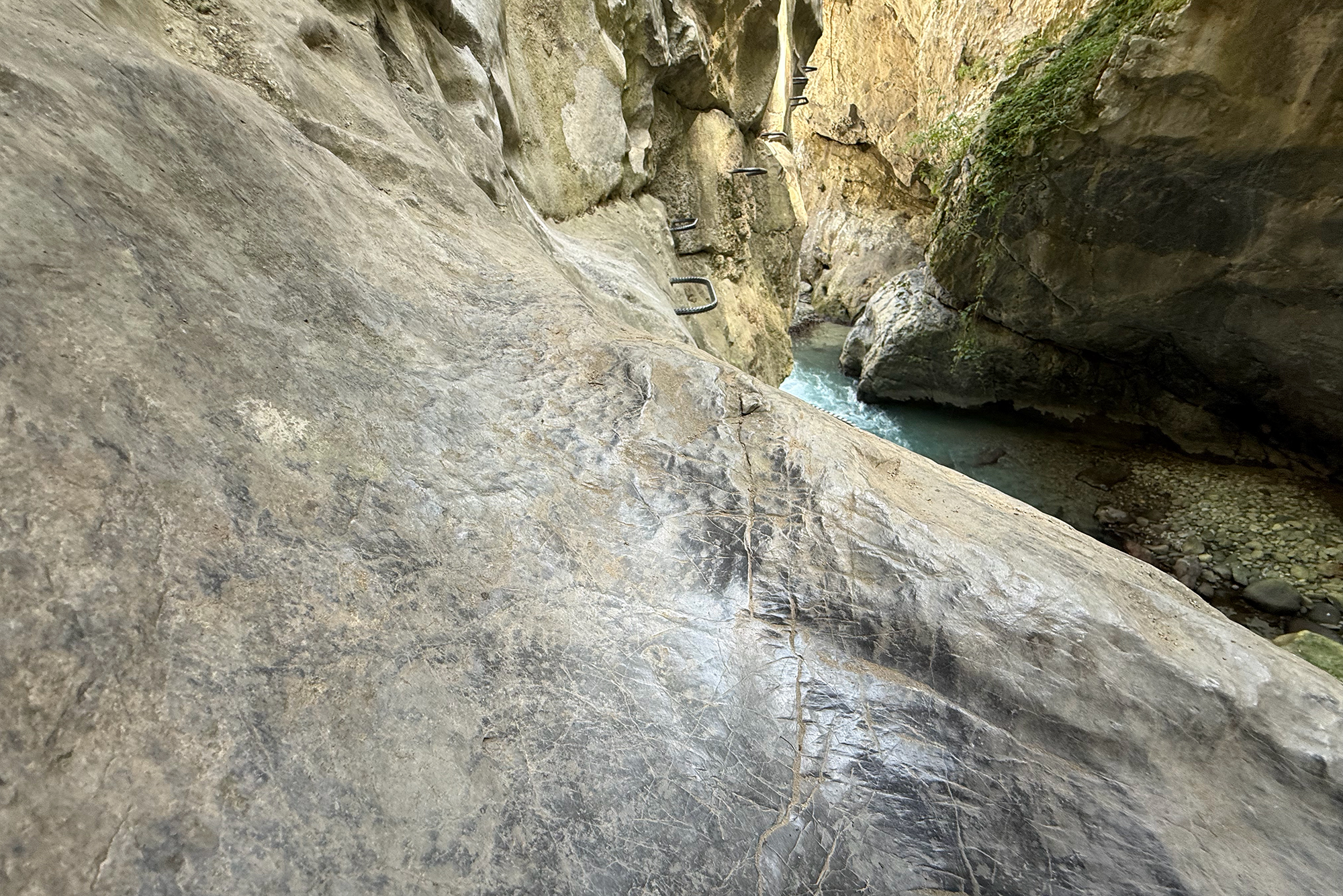 Rocky canyon wall with a metal climbing ladder attached to the rock, overlooking a narrow, blue-green river below.