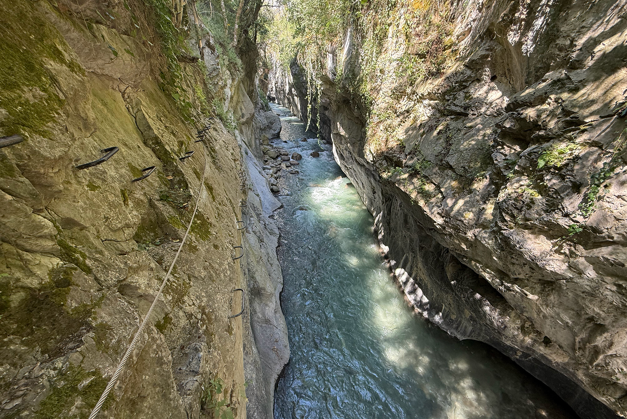 Steep rocky gorge with a fast-flowing turquoise river and a rope/ladder railing along the left wall for a canyon path