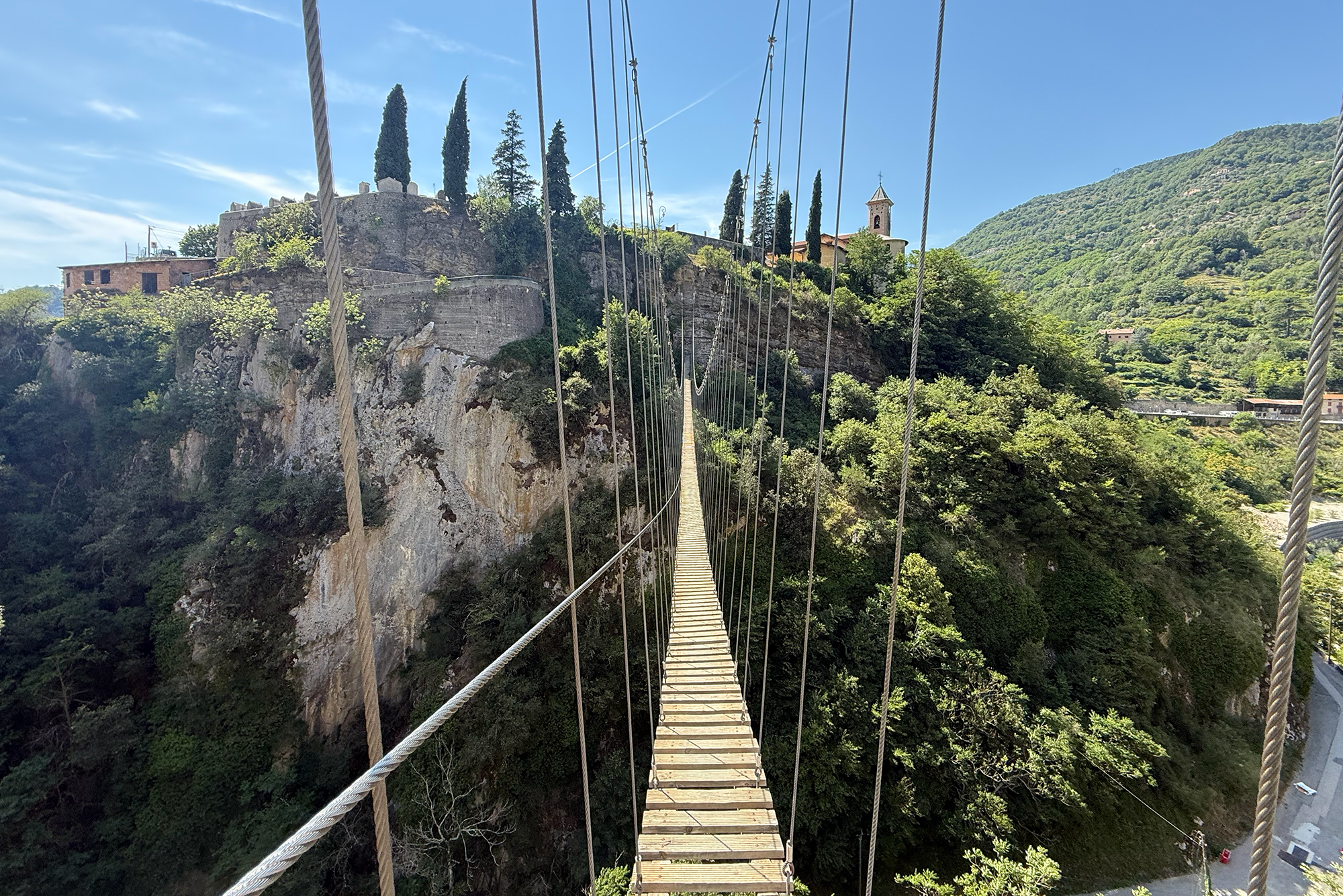 Long wooden suspension bridge crossing a deep gorge, stone fortress on the left and a church-topped hill to the right.