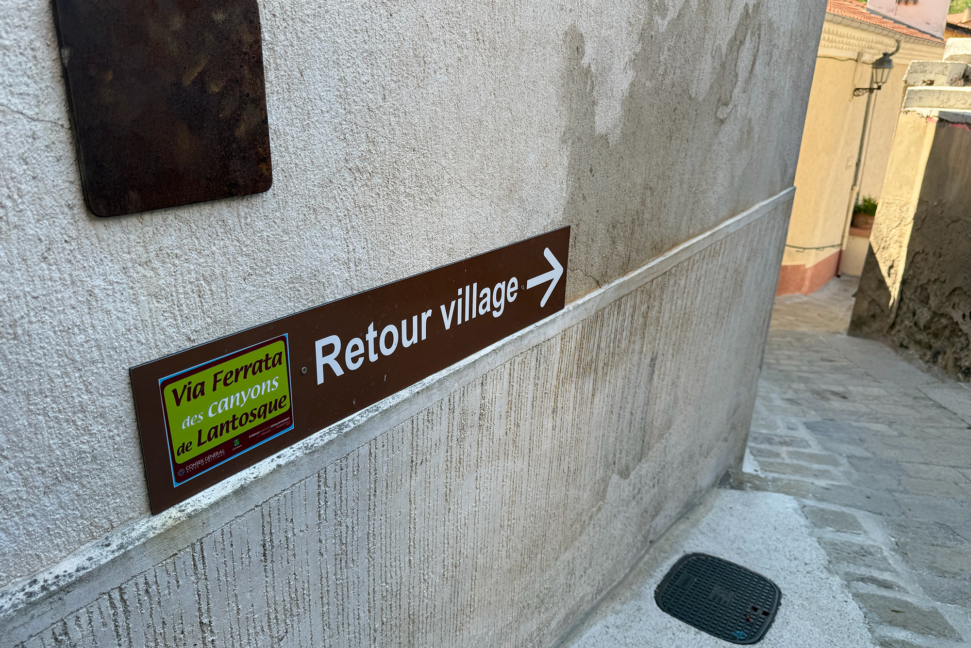 Brown directional sign reading 'Retour village' with a right arrow mounted on a textured exterior wall of a narrow alley, with a smaller colorful sign beneath it still visible.