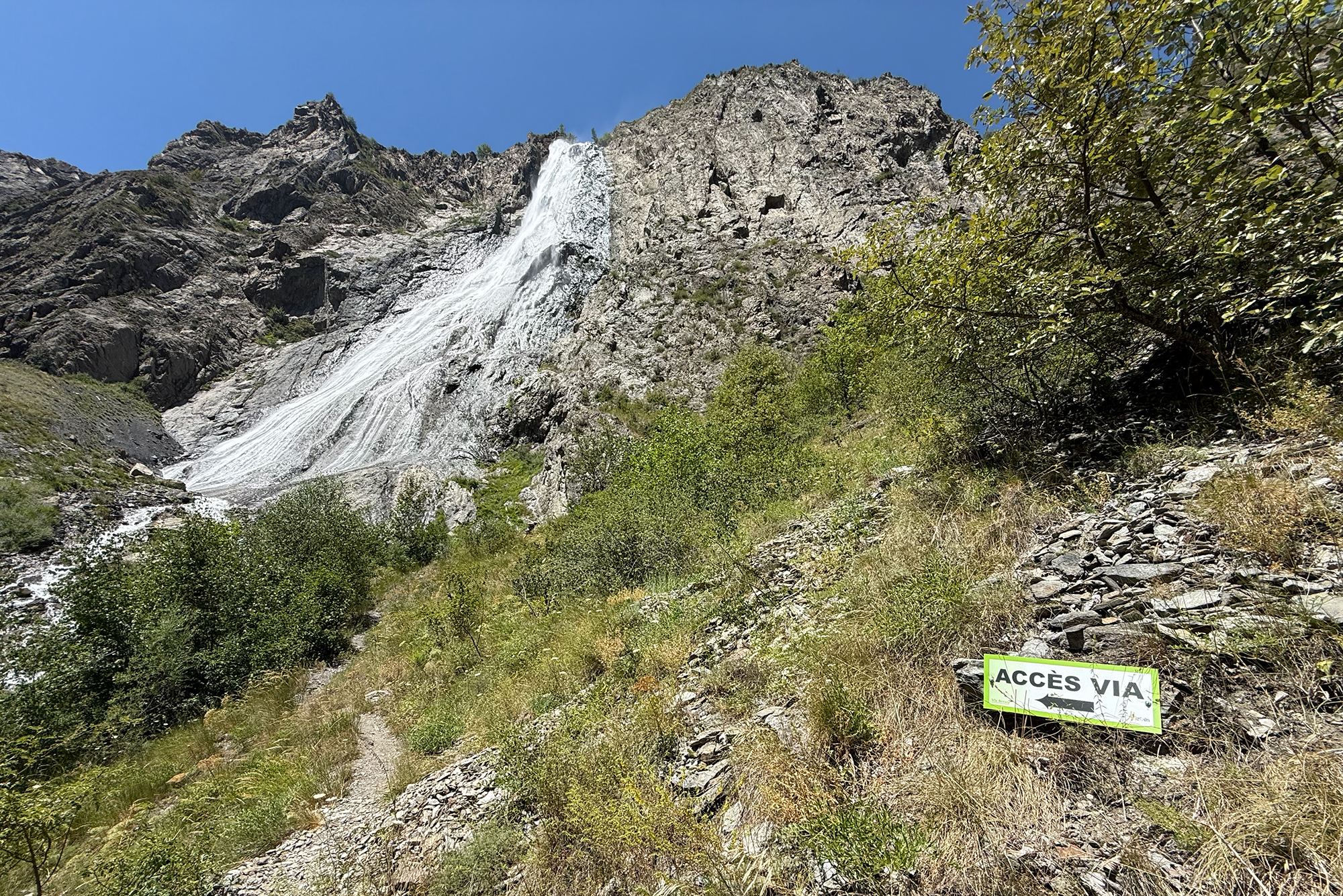Rugged rocky mountainside with a white waterfall-like runoff descending the cliff; foreground has dry grasses and shrubs under a clear blue sky.