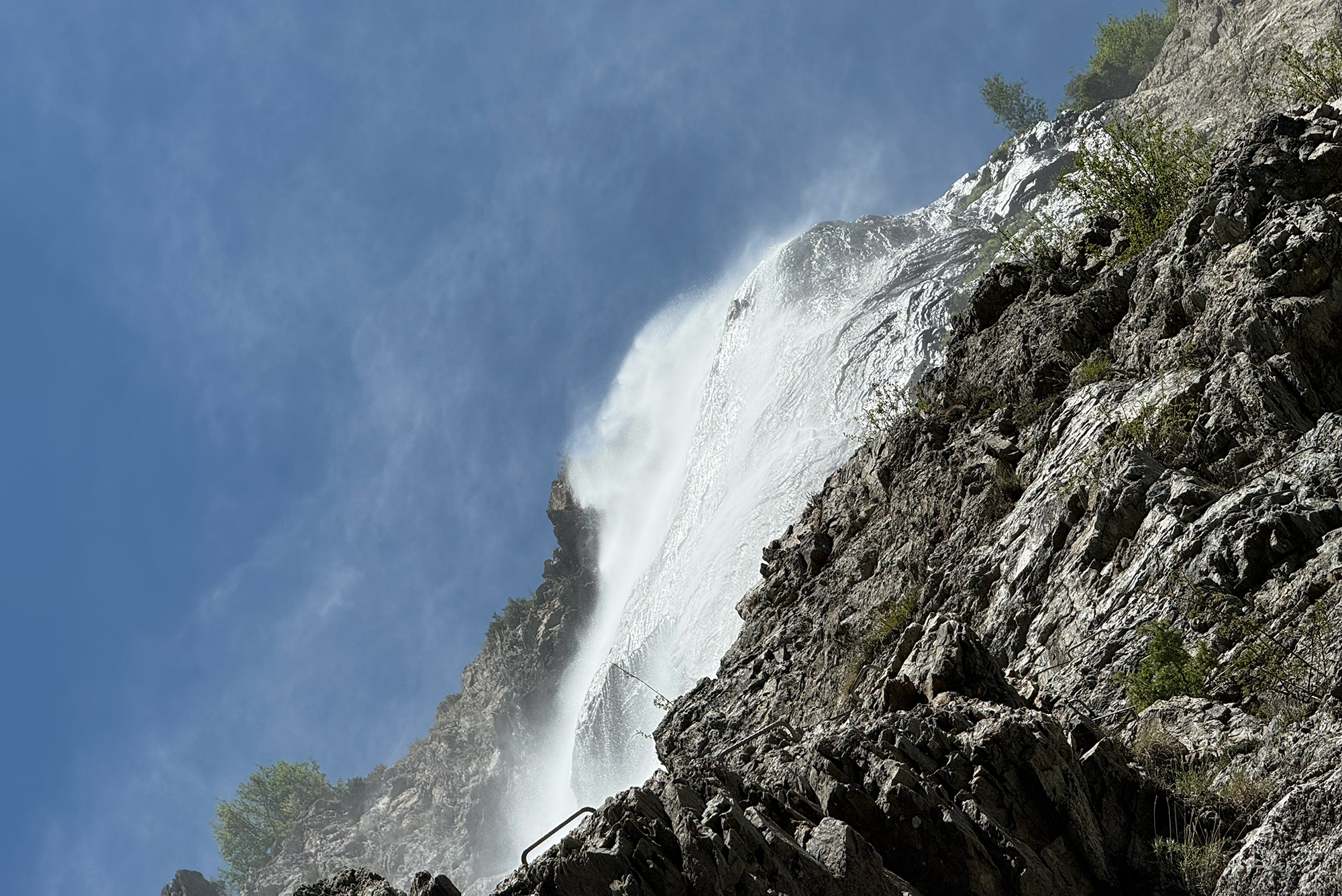 Waterfall cascading down a rugged rocky cliff with mist and a blue sky background.