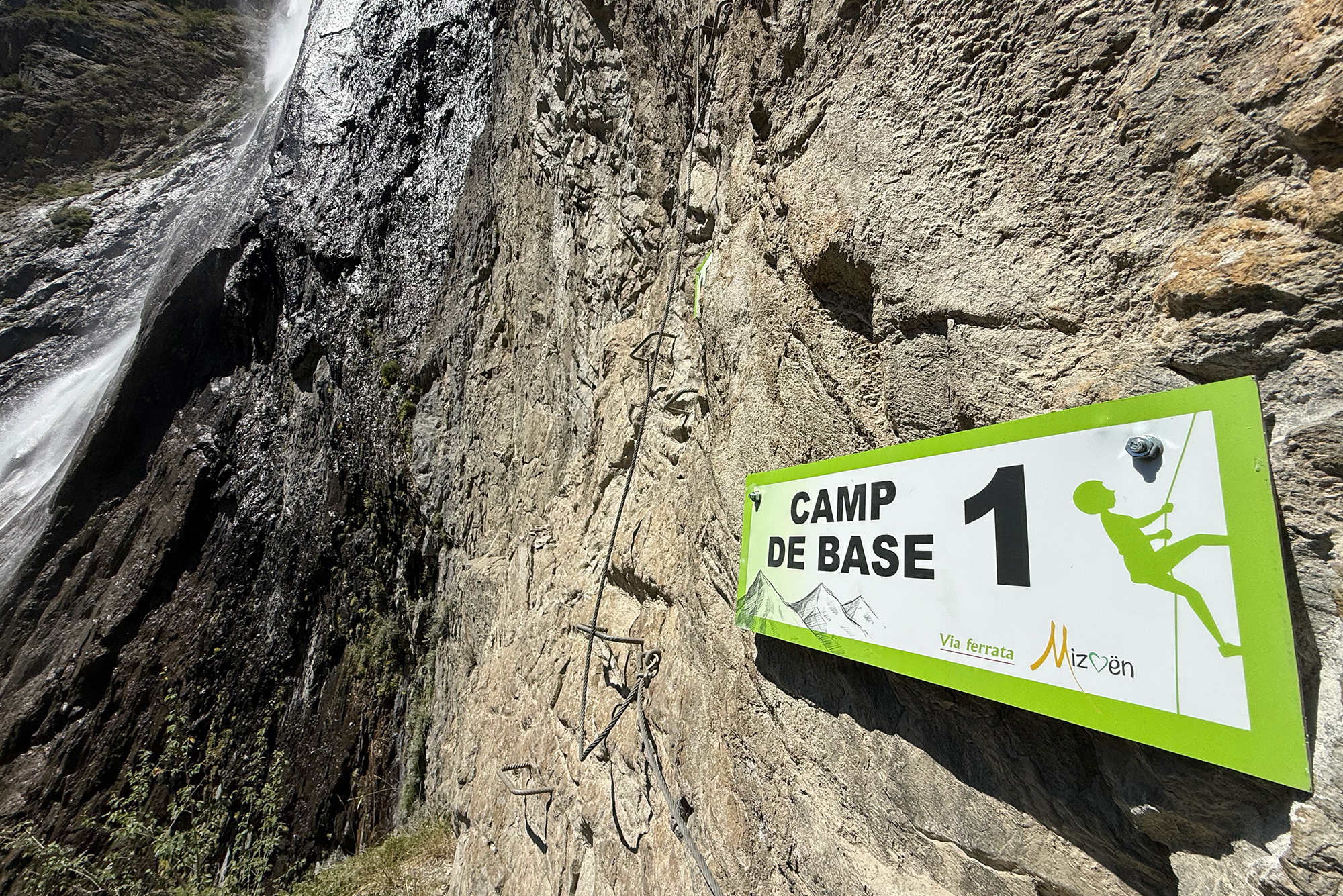 Rugged rock cliff with climbing ropes and a green 'Camp de Base 1' sign on the wall near a waterfall edge.