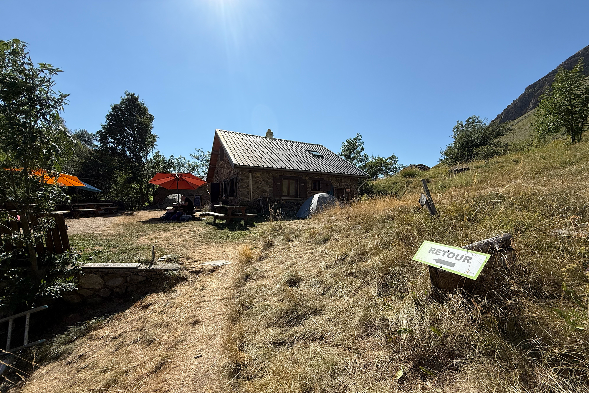 Sunny mountain retreat with a stone cabin, metal roof, and orange umbrellas, under a bright blue sky.