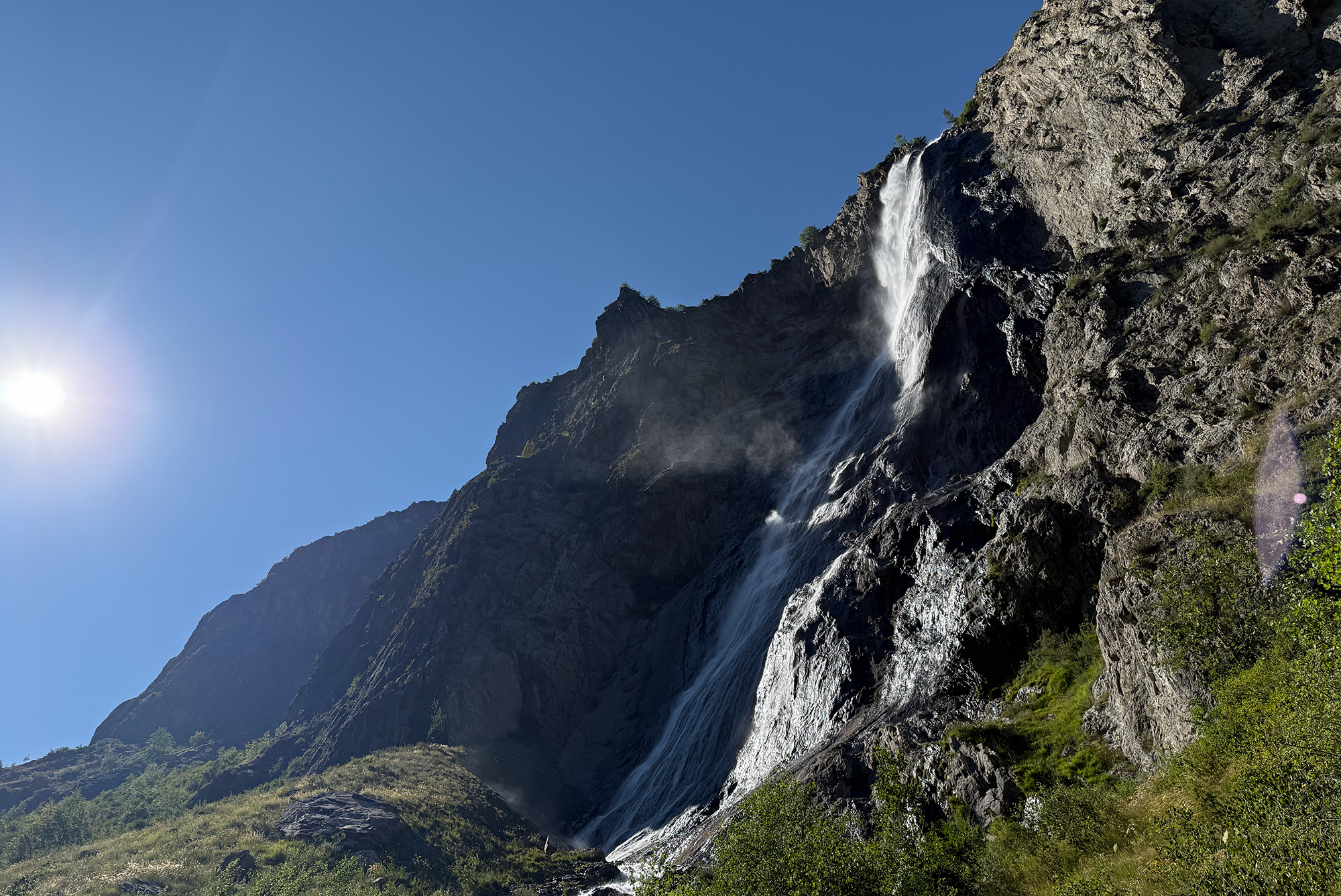 Waterfall cascading down a rocky cliff under a clear blue sky, with green vegetation at the base.