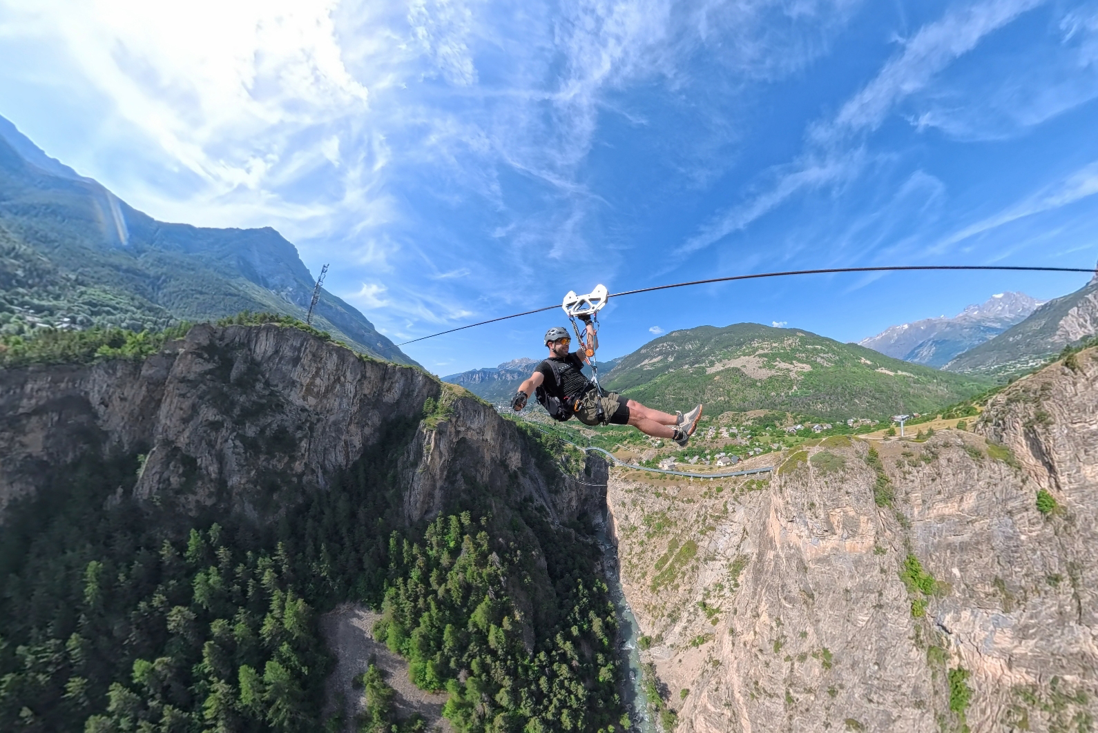 Person wearing helmet and harness ziplining across a deep canyon between rocky cliffs under a blue sky with mountains in the distance