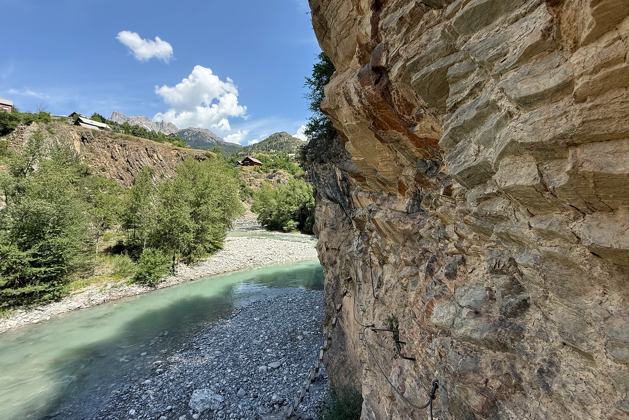 River canyon scene with a turquoise stream, rocky cliff on the right, trees and pebbled shore on the left, and mountains in the distance under a blue sky.