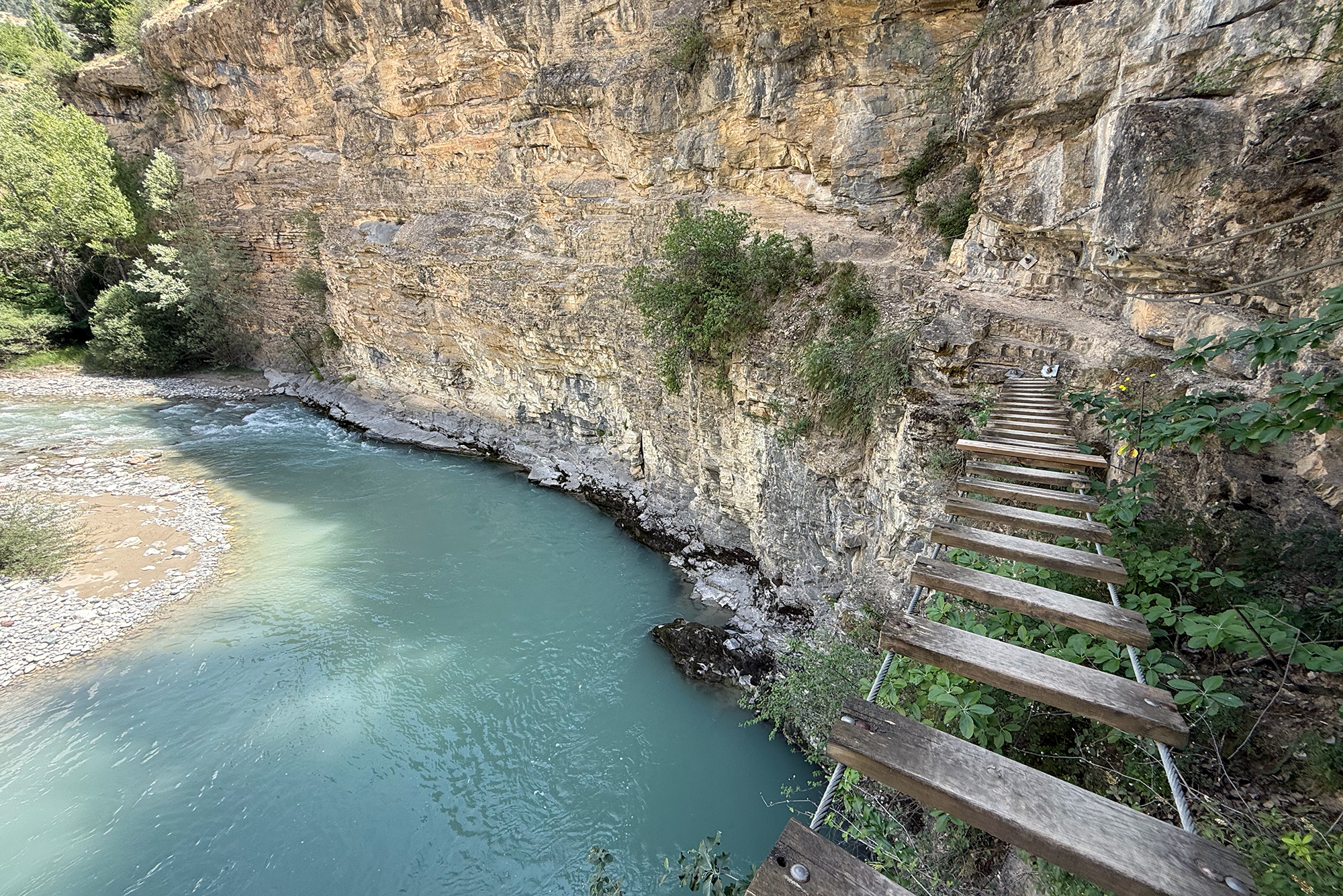 Rocky cliff faces the turquoise river; a weathered wooden boardwalk runs along the right edge.