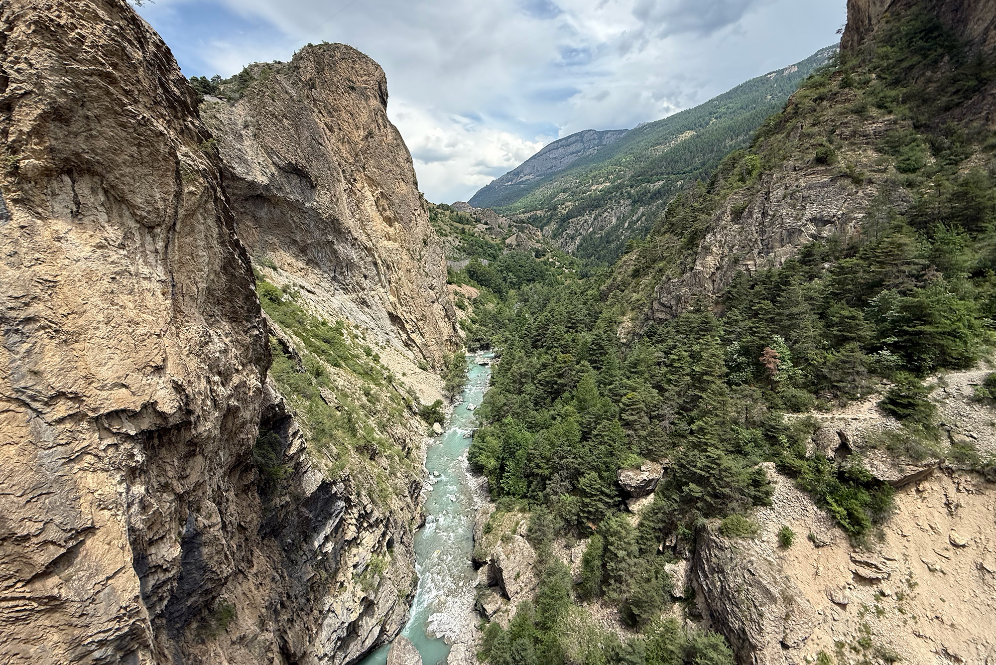 Steep rocky canyon with a turquoise river cutting through, surrounded by pine trees and rugged cliffs under a cloudy sky from a high vantage point