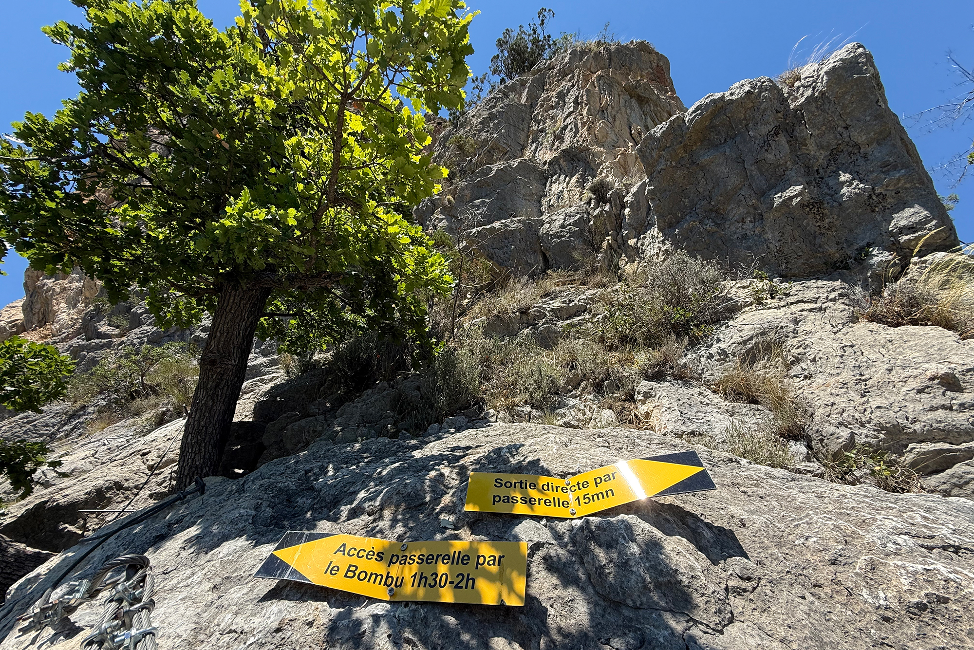 Rocky slope with a green tree in front; bright yellow trail signs in French warn about a footbridge route (15 minutes).