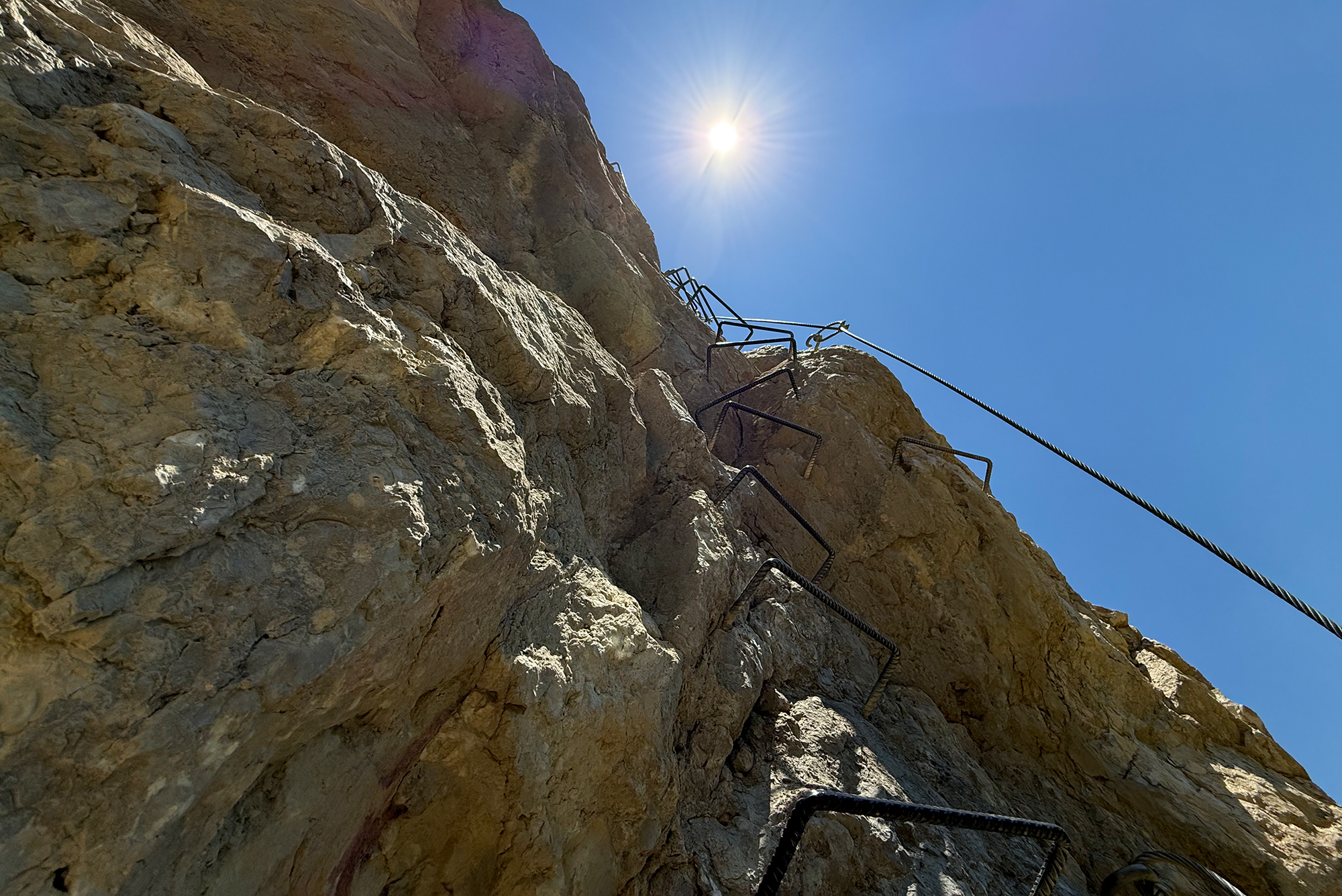 Rocky cliff with metal rungs attached for climbing and a safety rope against a clear blue sky.