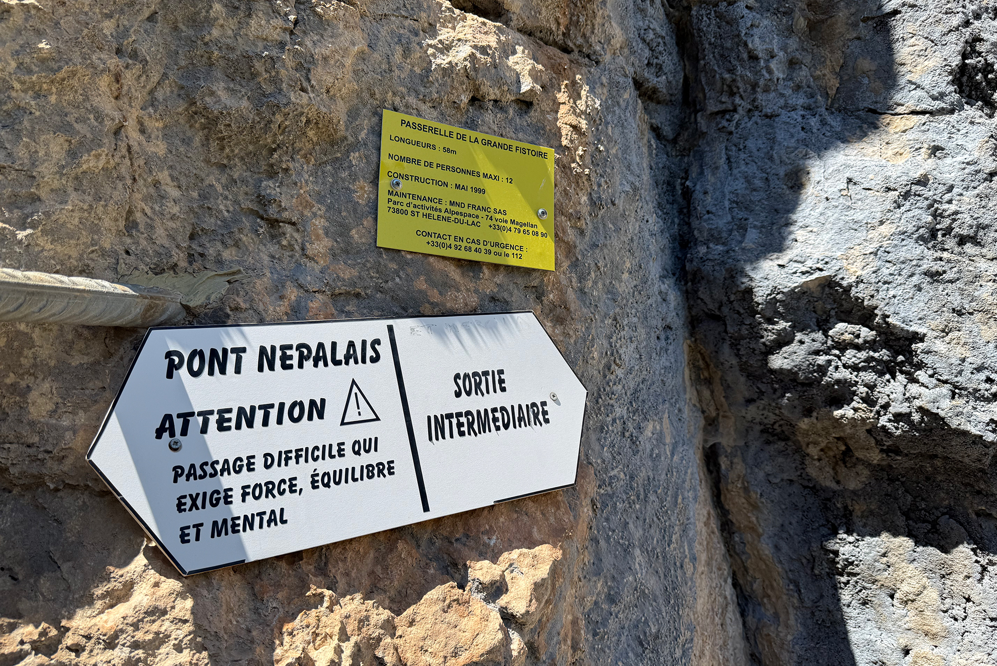 Rocky cliff with two signs: a yellow information plaque and a white warning sign mounted on the rock wall.
