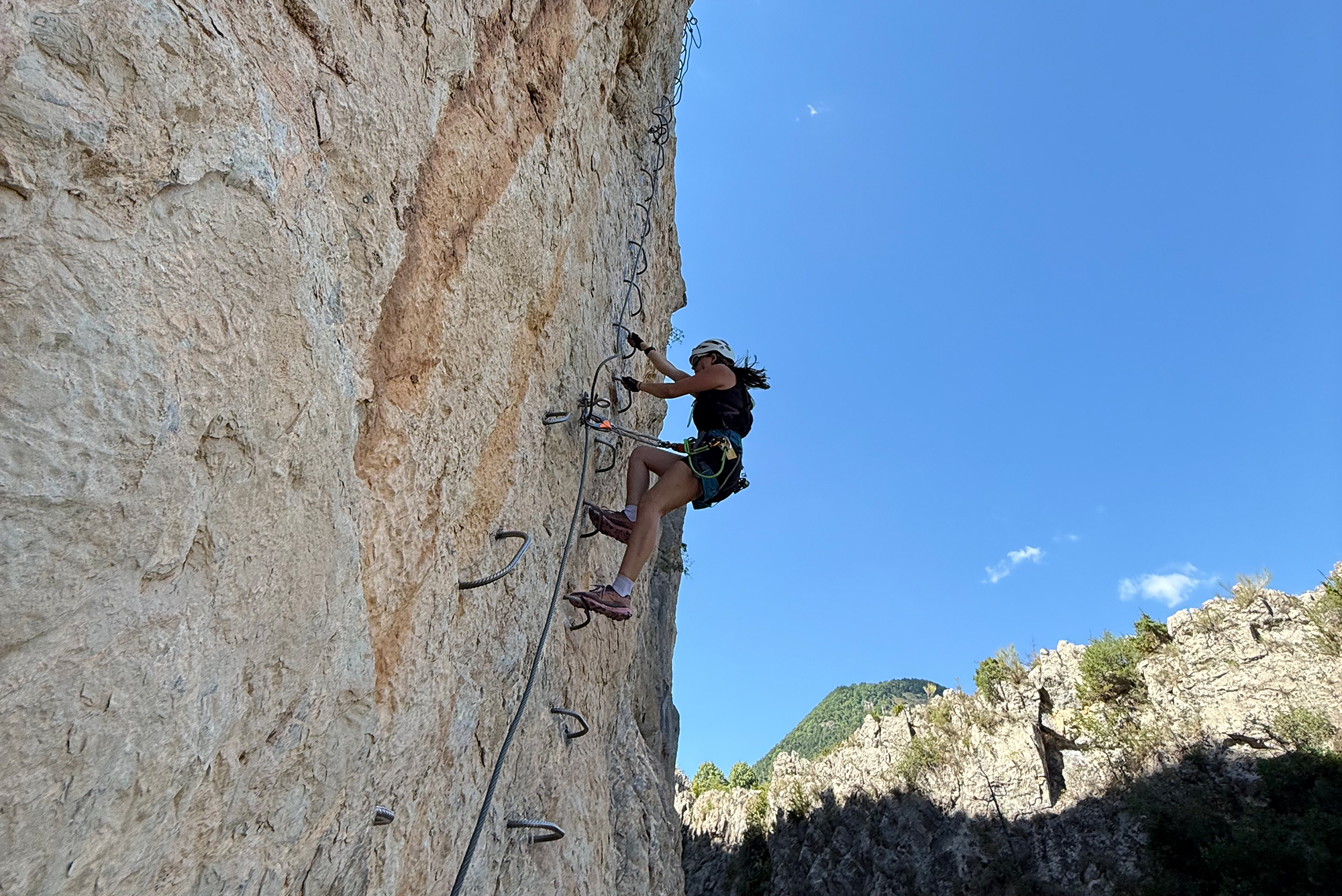 Climber wearing helmet and harness ascends a steep rocky cliff with a rope against a clear blue sky