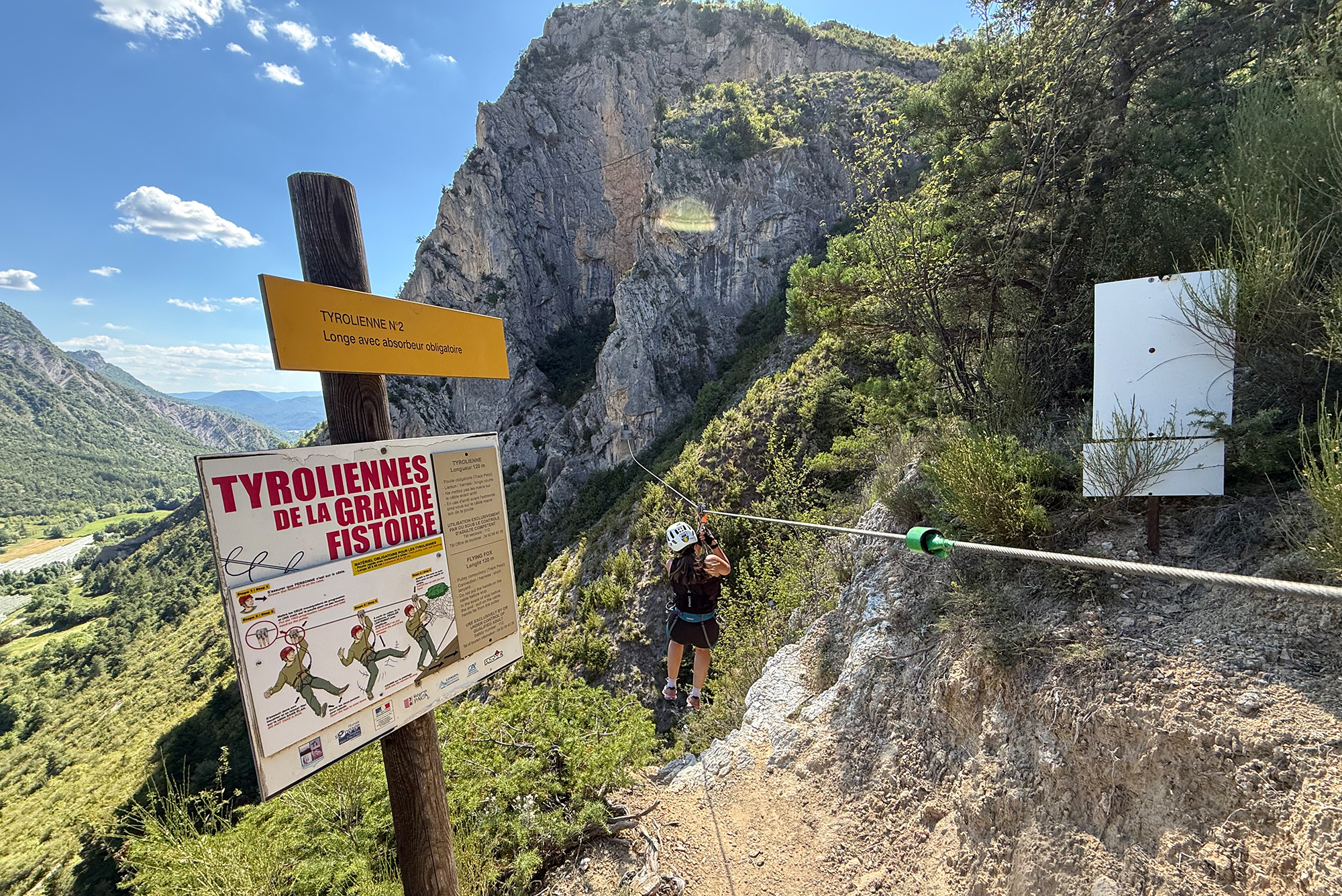 Trail sign area in a rocky gorge with a yellow sign reading Tyroliennes N°2 and a person in harness preparing for a zipline ride below the cliff edge.