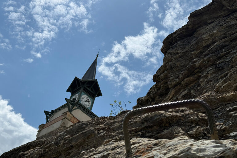 Clock tower with a pointed spire peeking above a rocky wall, set against a blue sky with white clouds