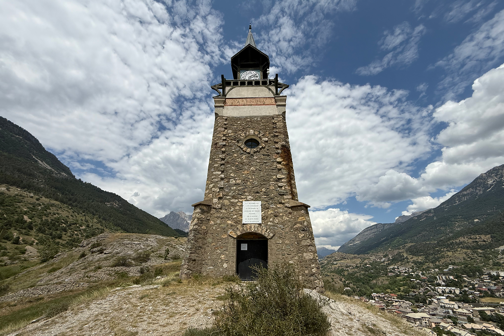 Stone clock tower on a rocky base, with blue sky and white clouds, overlooking a mountain valley and village below