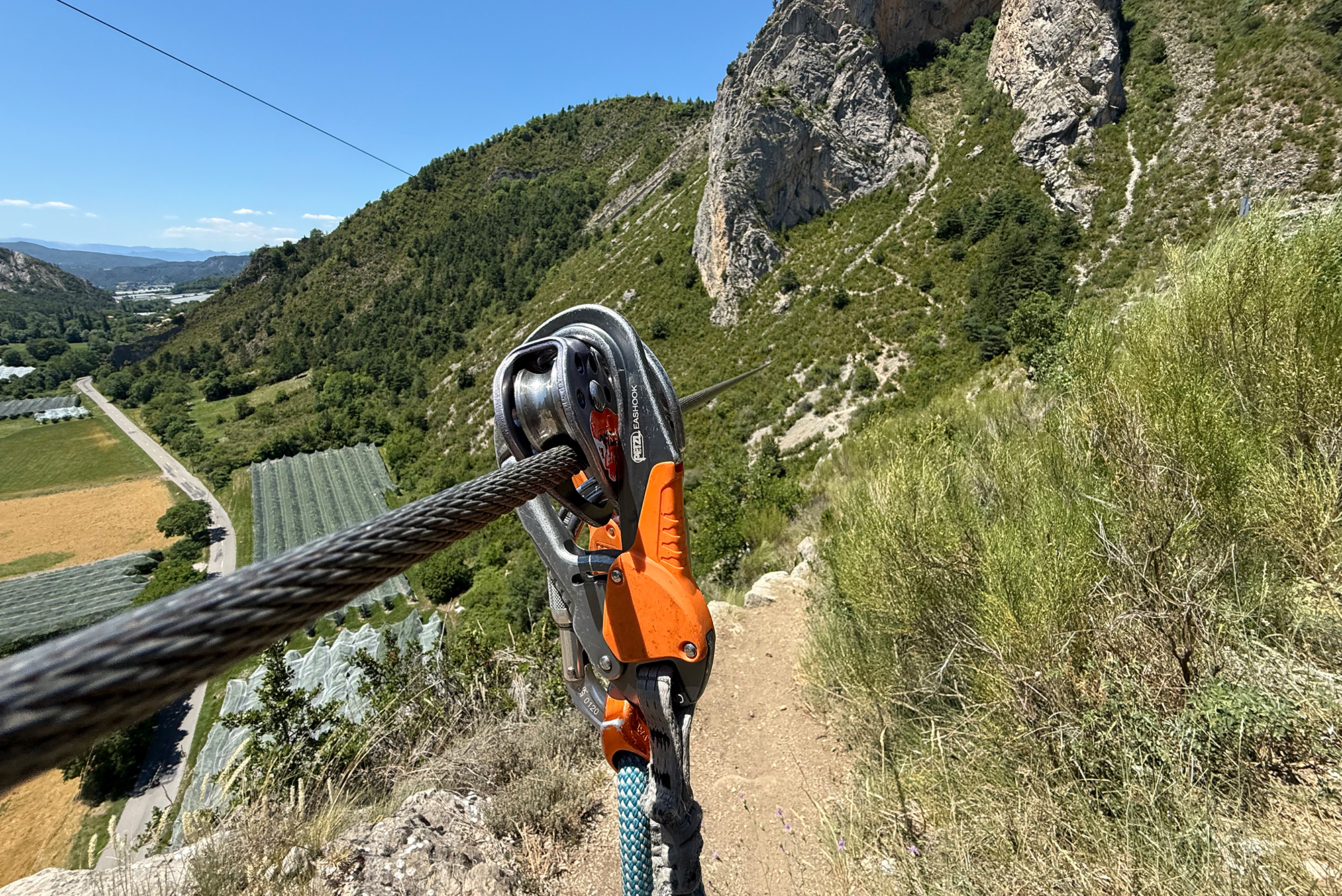Rope and belay device secured on a mountain trail, with rocky slopes and a valley visible in the distance behind.