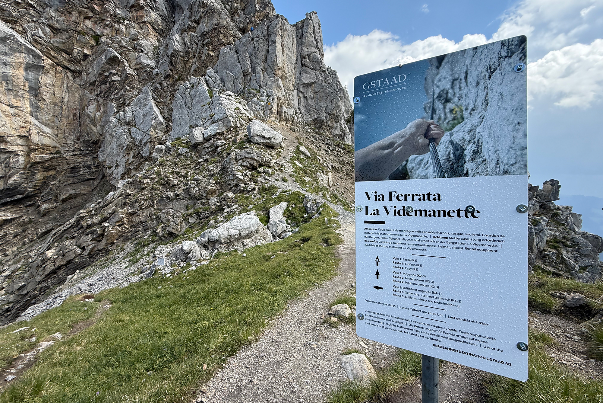 Rugged alpine rock wall with a large sign reading 'Via Ferrata La Videmanette' and a climber holding a rope on the rock face.