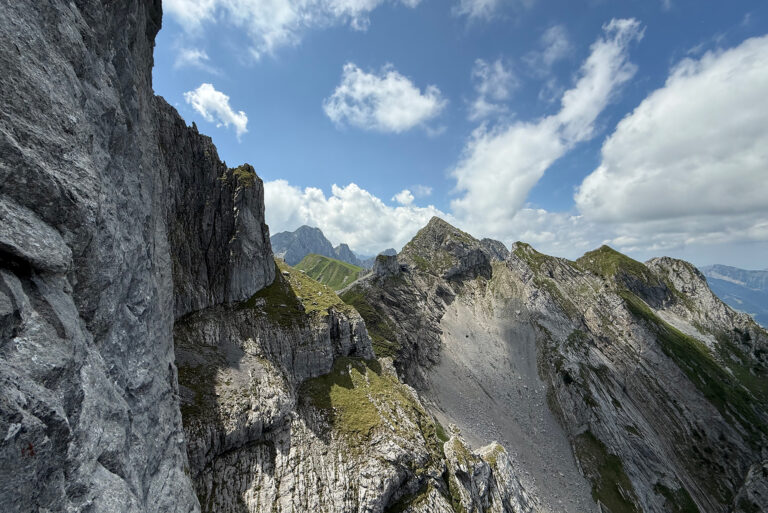 Rugged mountain landscape with jagged gray rock cliffs, green slopes, and a blue sky with white clouds, mid-day light.