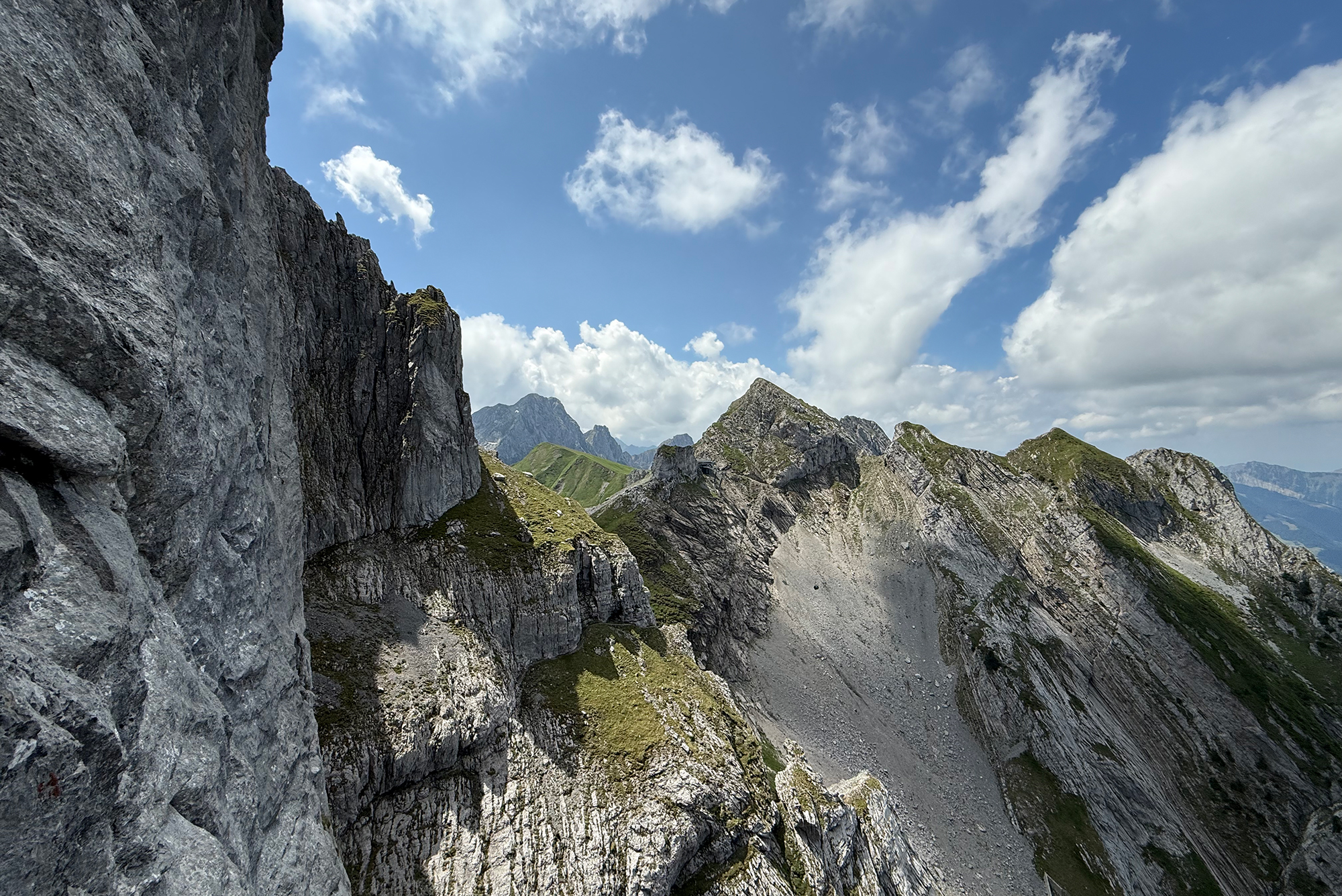 Rugged mountain landscape with jagged gray rock cliffs, green slopes, and a blue sky with white clouds, mid-day light.