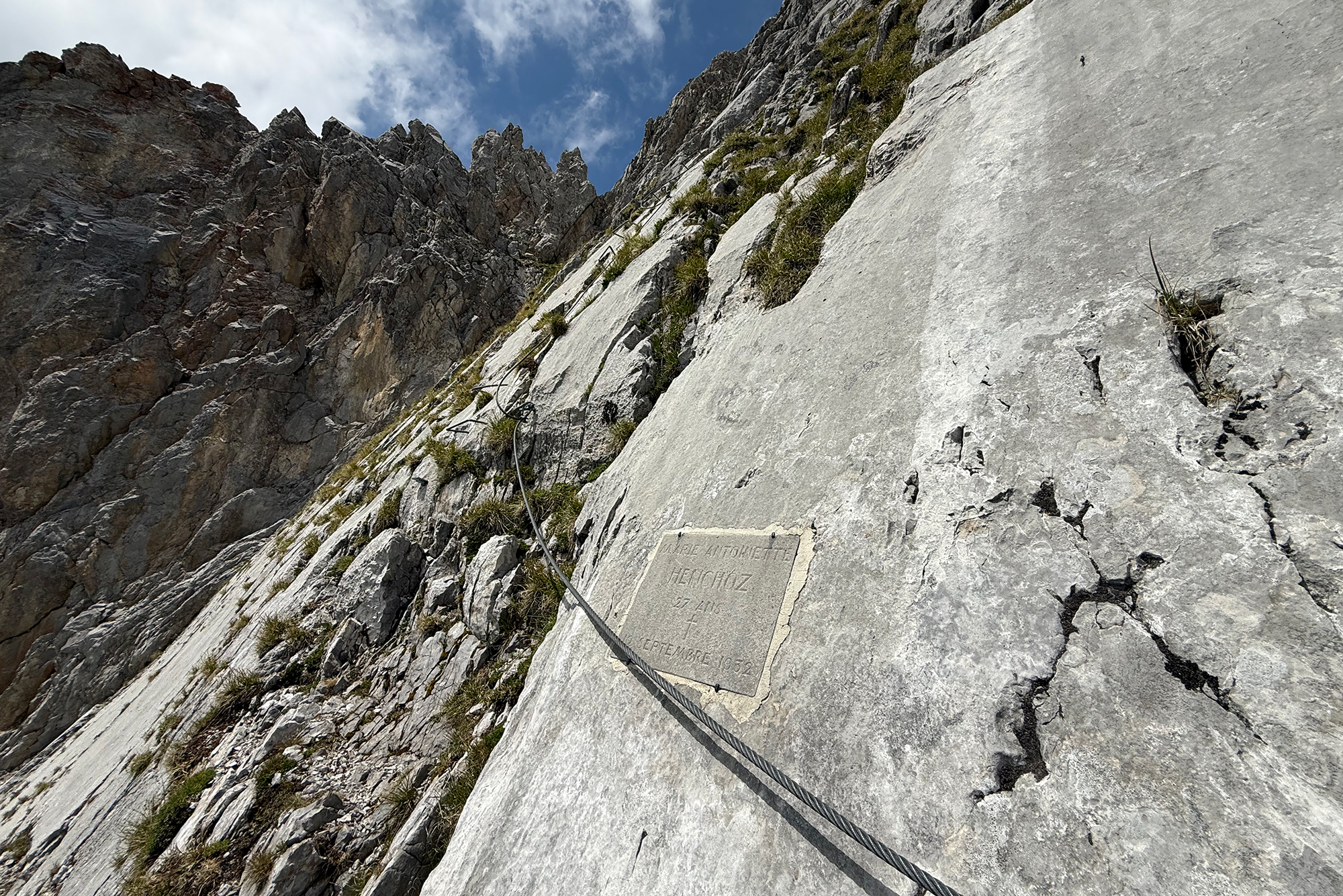Steep rocky cliff with a metal safety cable along a via ferrata route and sparse vegetation under a blue sky.