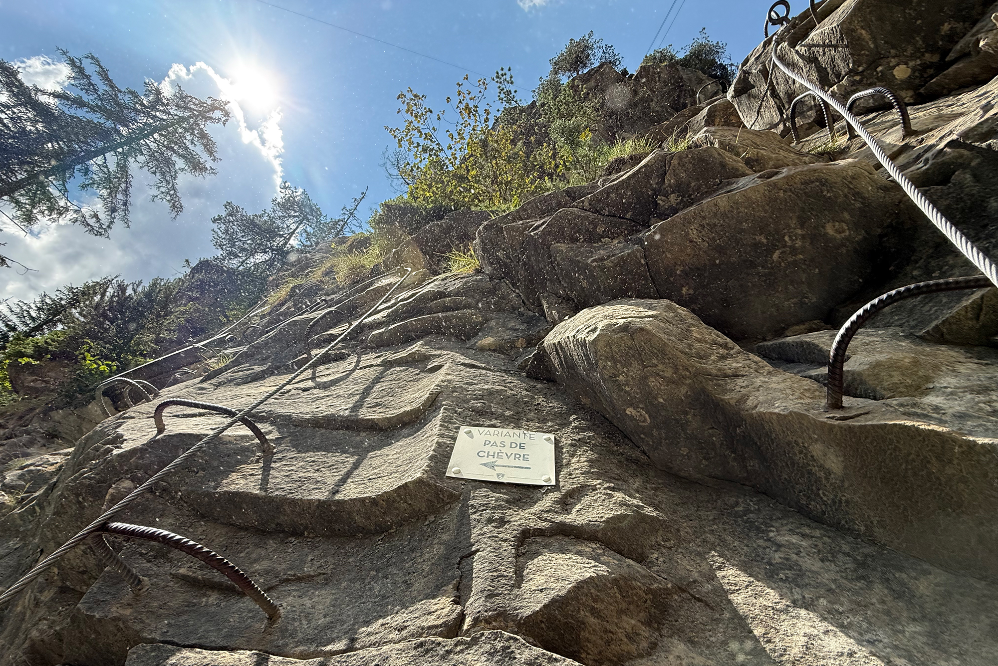 Rocky cliff with steel climbing cables and loops, sun over a blue sky and trees in the background; small sign reads 'Variante pas de chèvre'