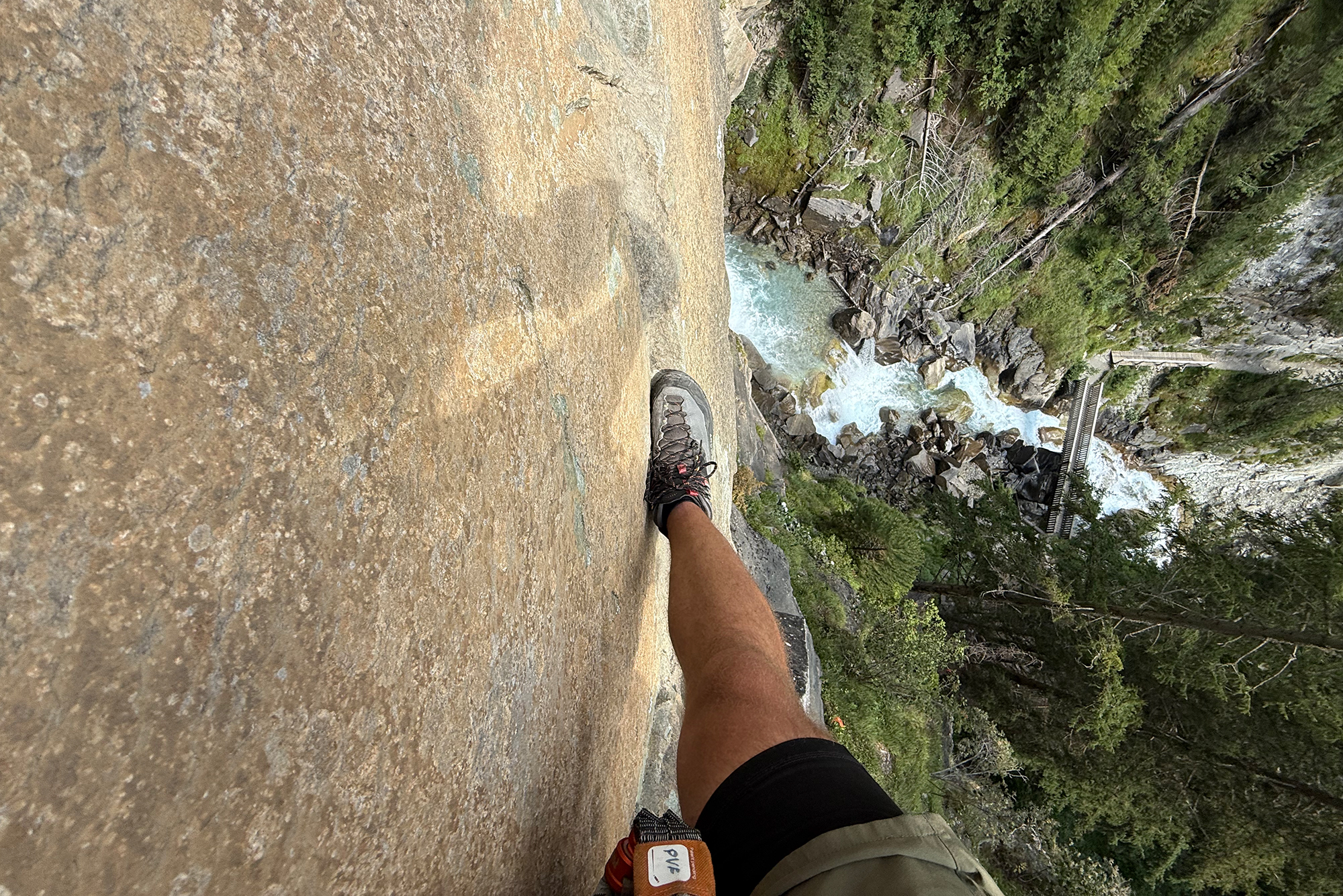 Person wearing hiking boots stands on a steep rock ledge overlooking a turquoise river and forested gorge below.