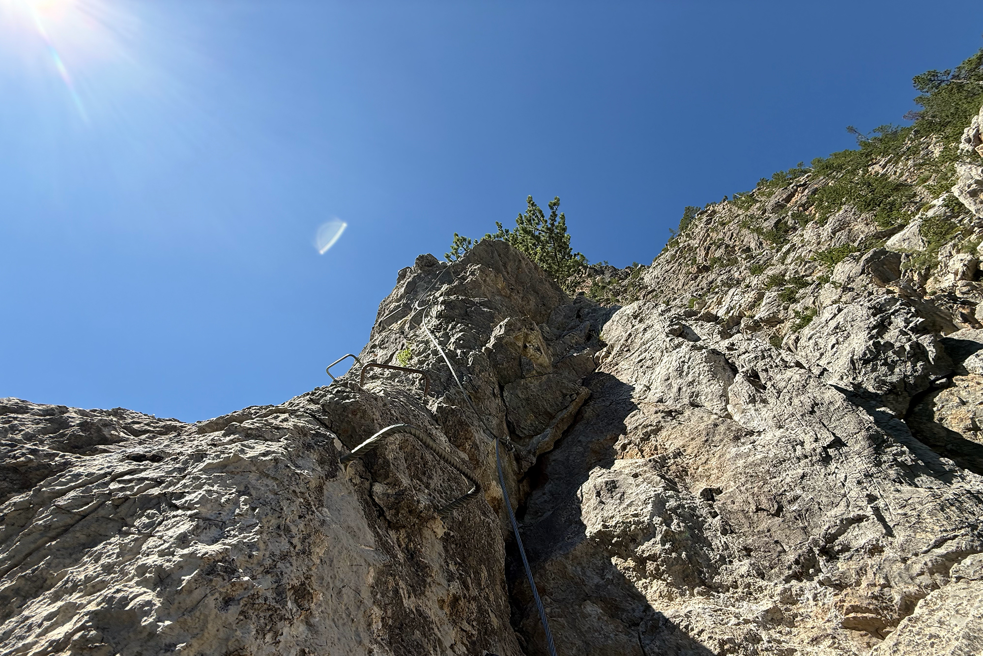 Steep rocky cliff with a climbing rope running up the crevice against a bright blue sky and sun flare.