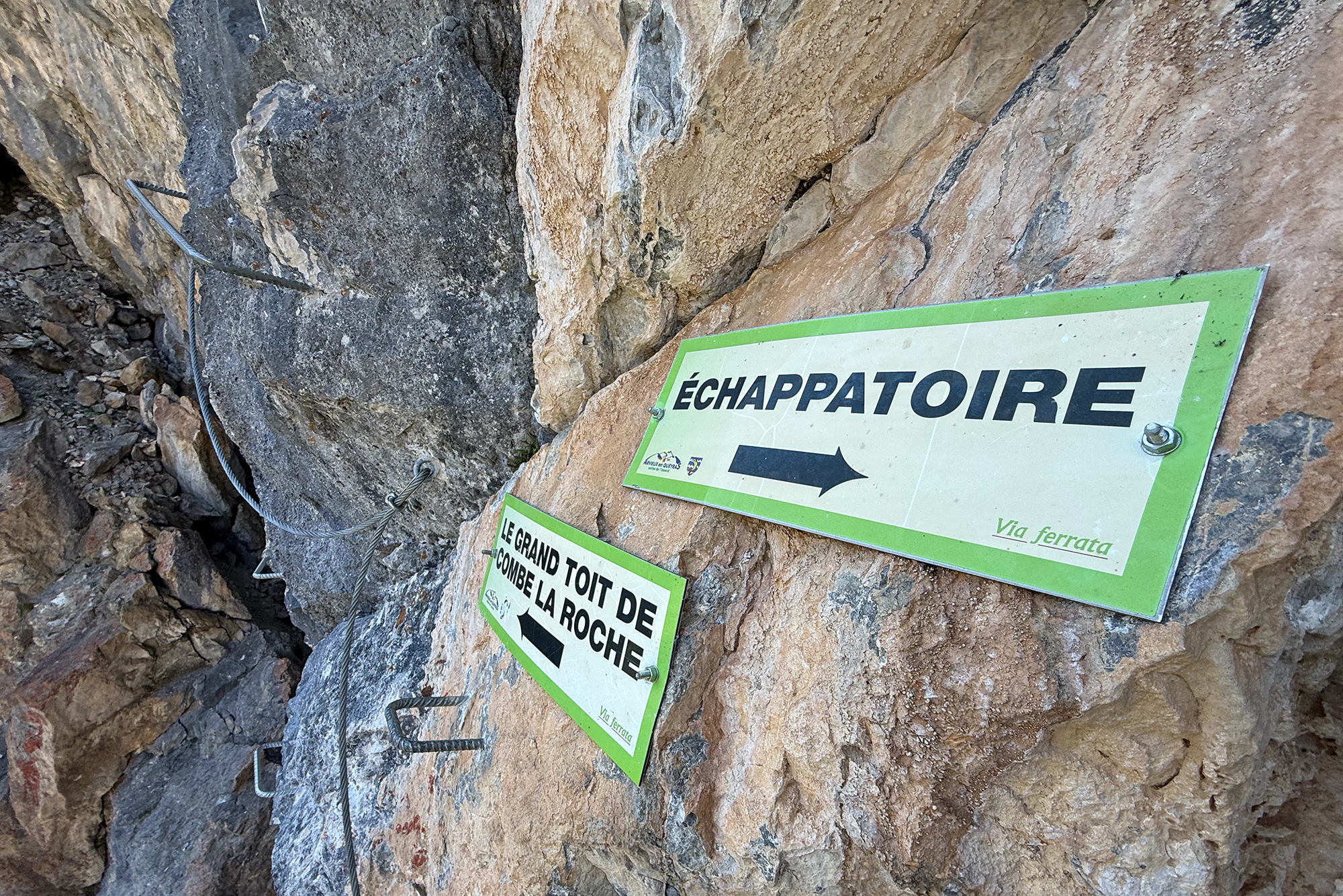 Rock cliff with metal safety cables and green-framed signs in French, indicating a via ferrata route ahead.